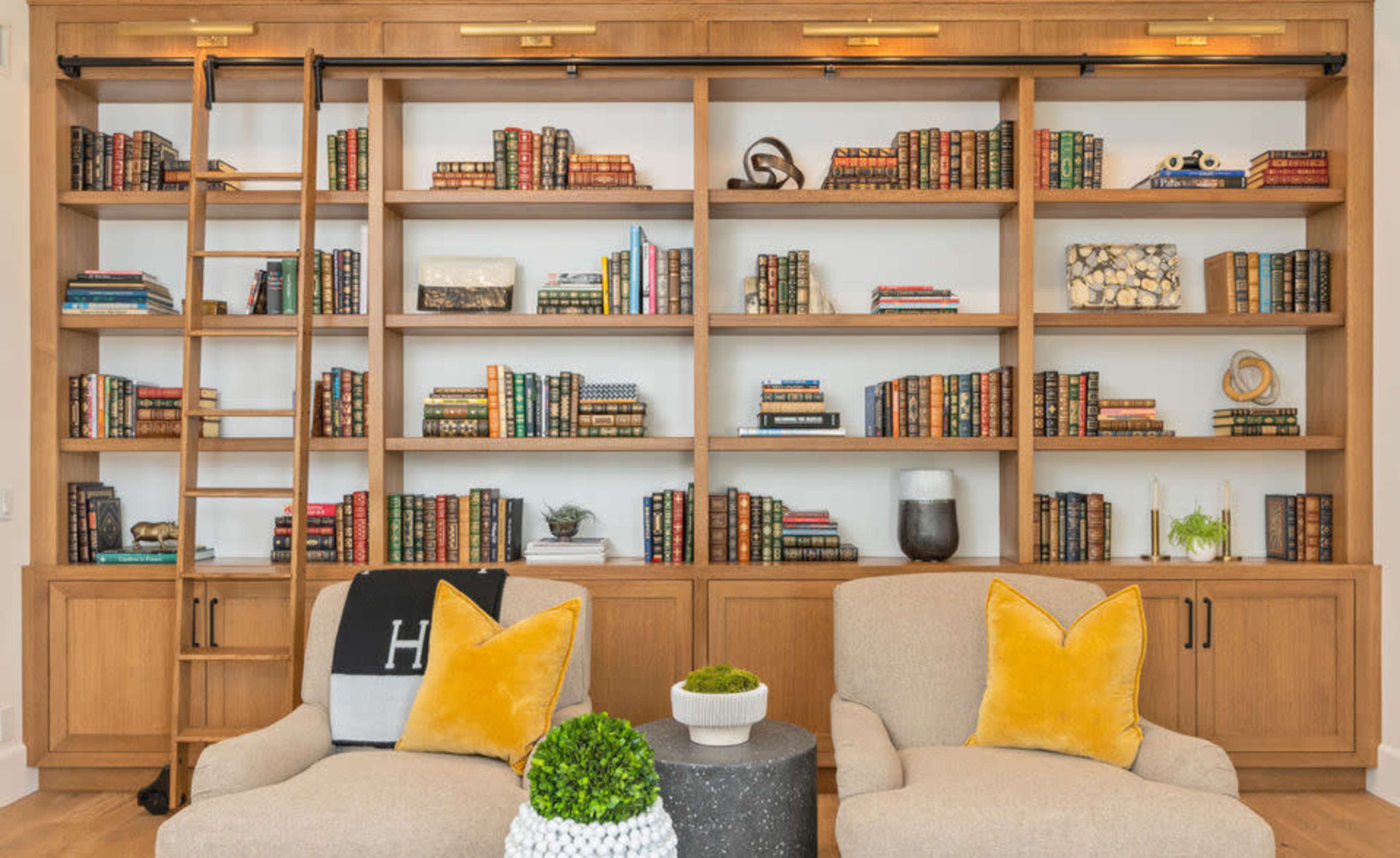 A wooden shelving unit filled with neatly arranged books and decorative items, flanked by two upholstered chairs and a ladder.