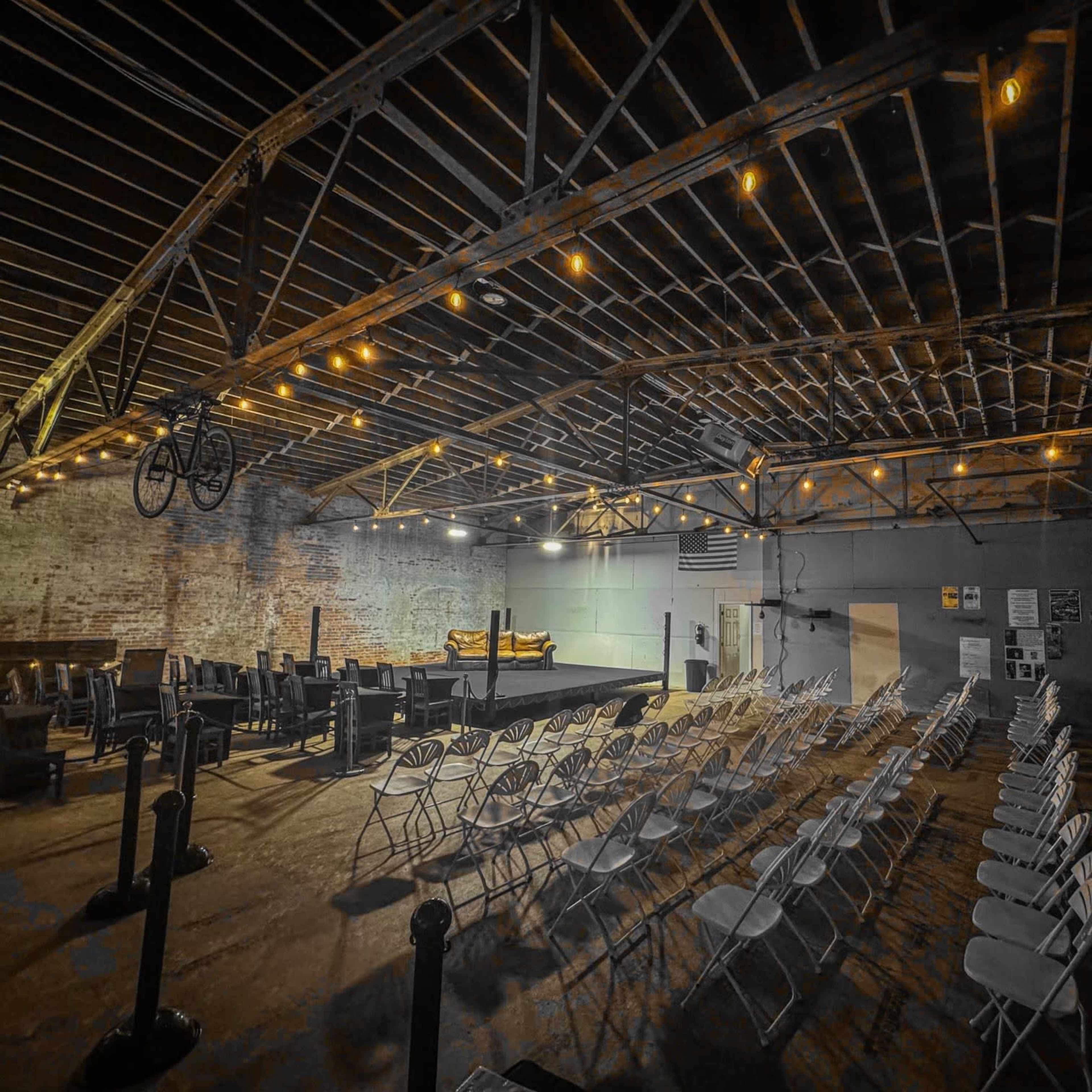 A spacious venue features rows of white chairs facing a performance stage under rustic wooden beams and overhead string lights.