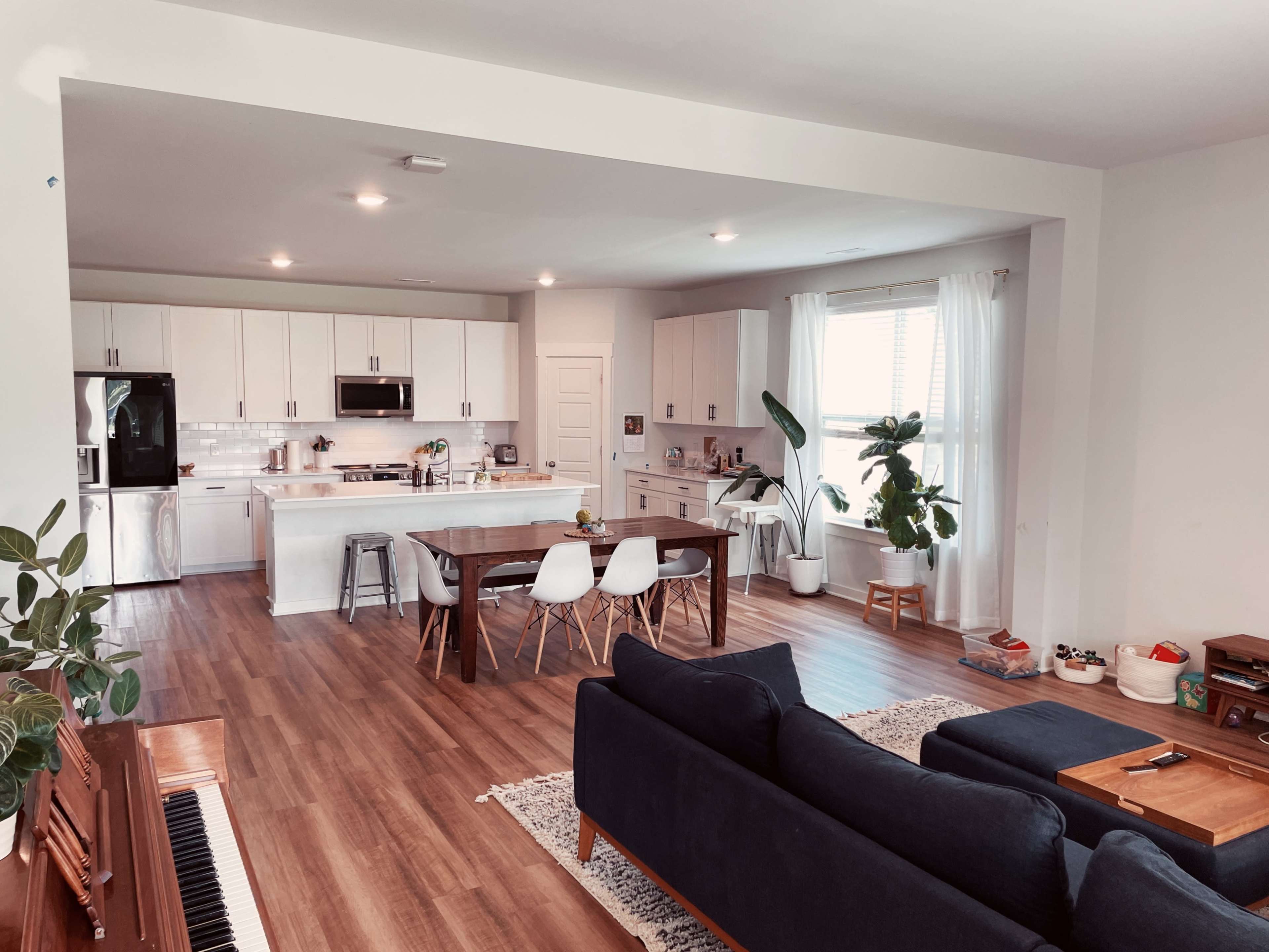 The image shows a modern open-concept kitchen and dining area with a wooden table, white chairs, and a sofa in a well-lit space.