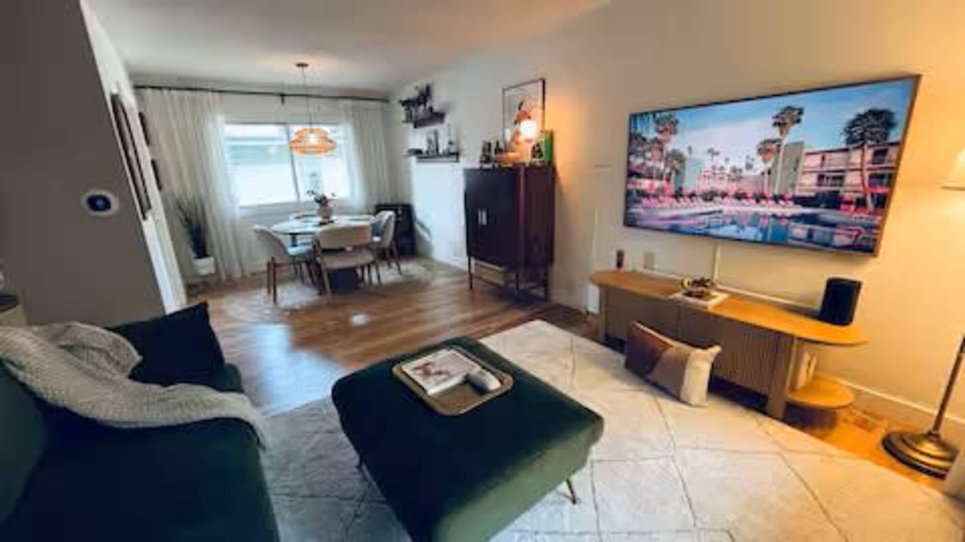 The image shows a cozy living room with a green sofa, a coffee table, a large television mounted on the wall, and a dining area visible in the background.
