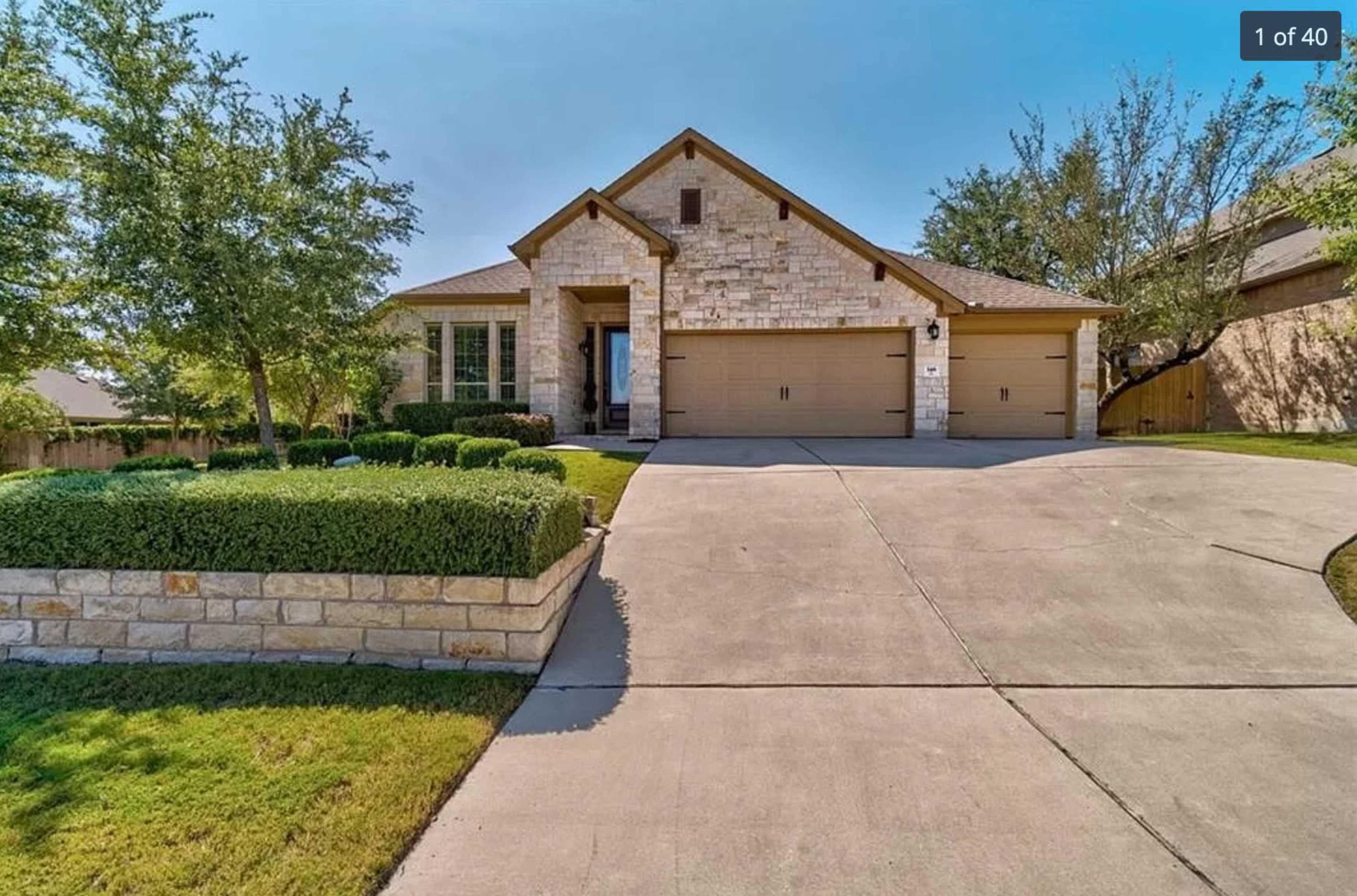 A single-story stone house with a two-car garage sits on a large concrete driveway surrounded by neatly trimmed shrubs and trees.