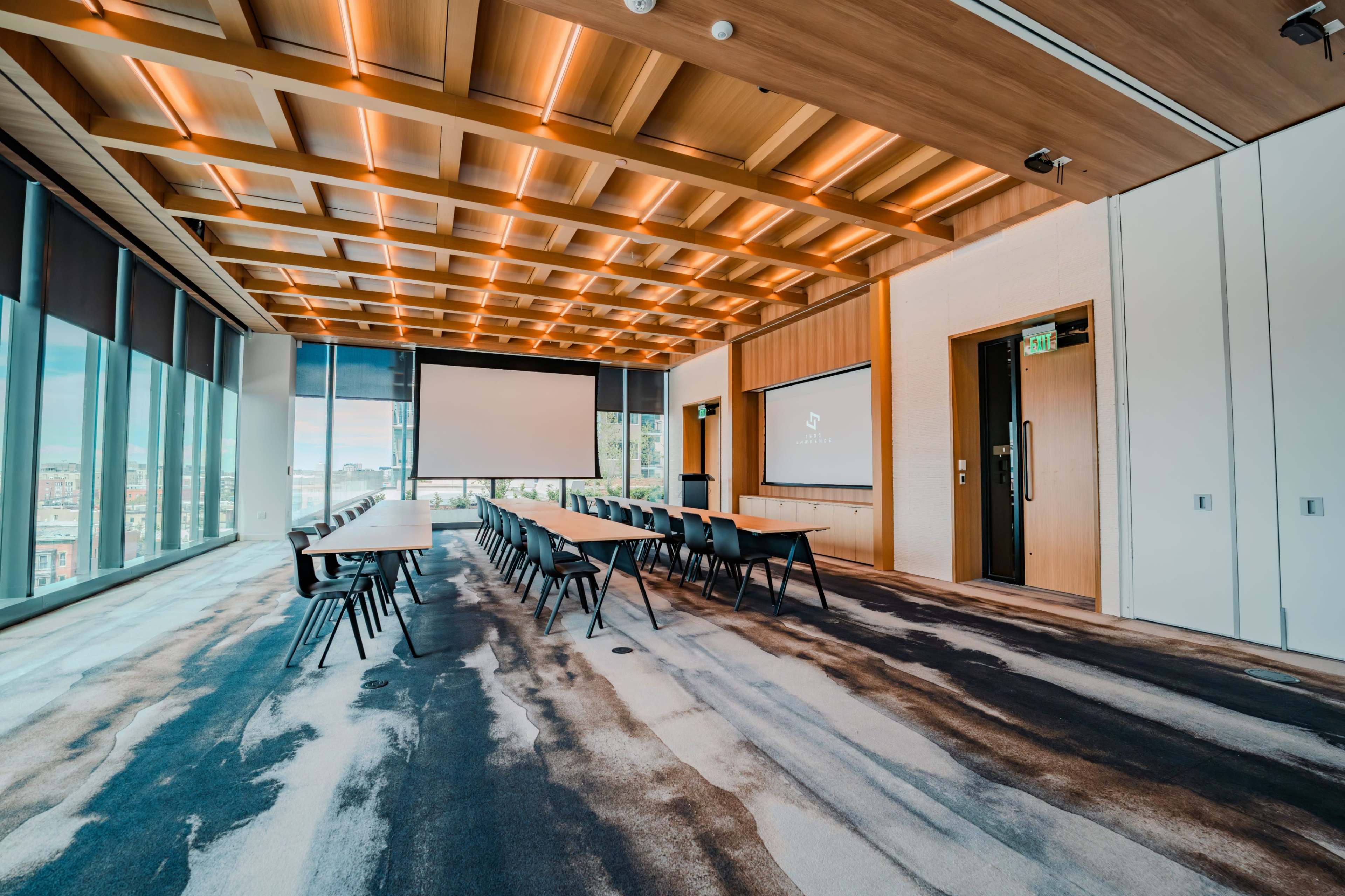 A modern conference room features a long table with chairs and large windows, illuminated by overhead lighting and equipped with presentation screens.