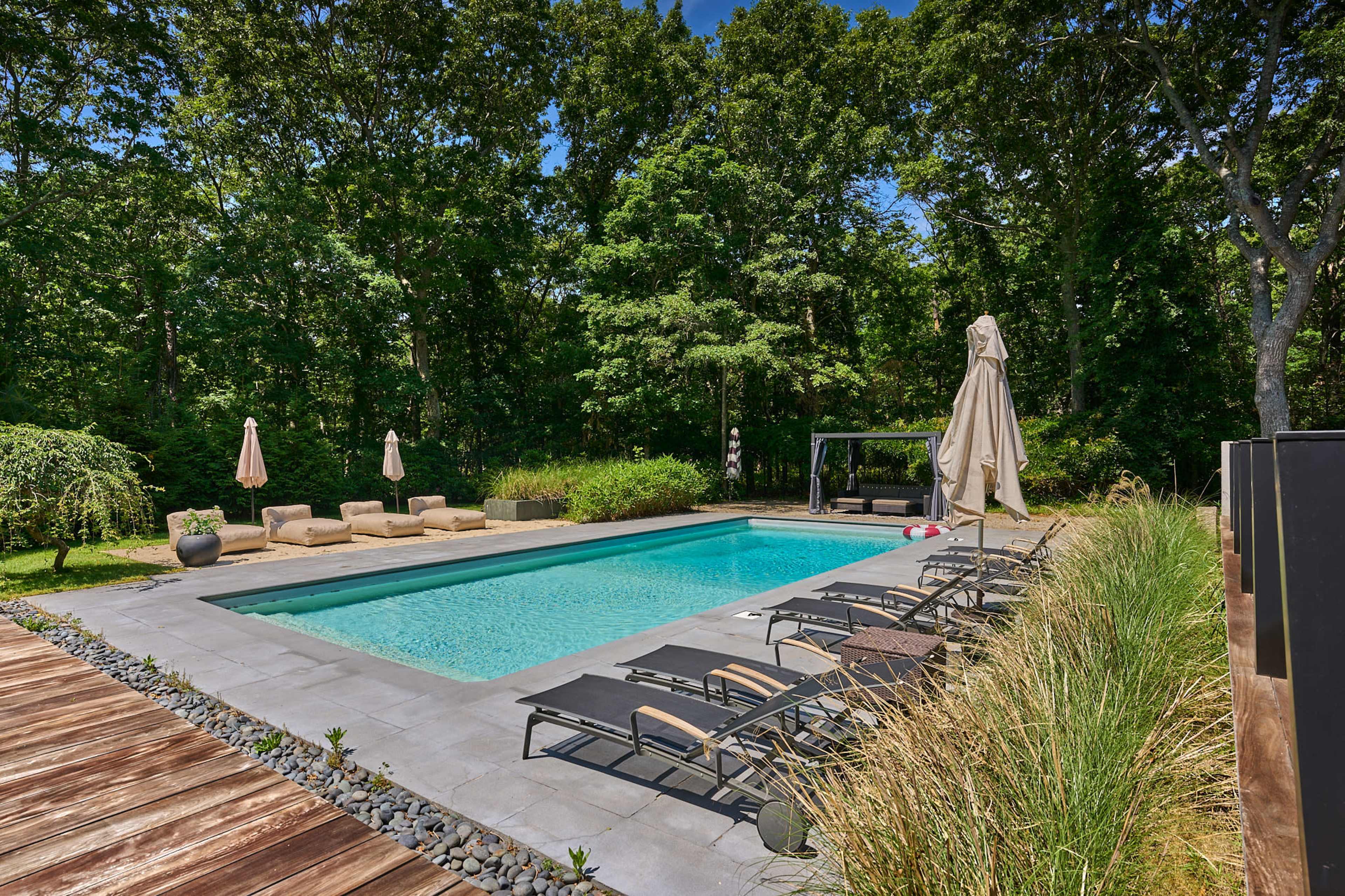 The image shows a modern swimming pool surrounded by green trees, with lounge chairs and a shaded seating area nearby.