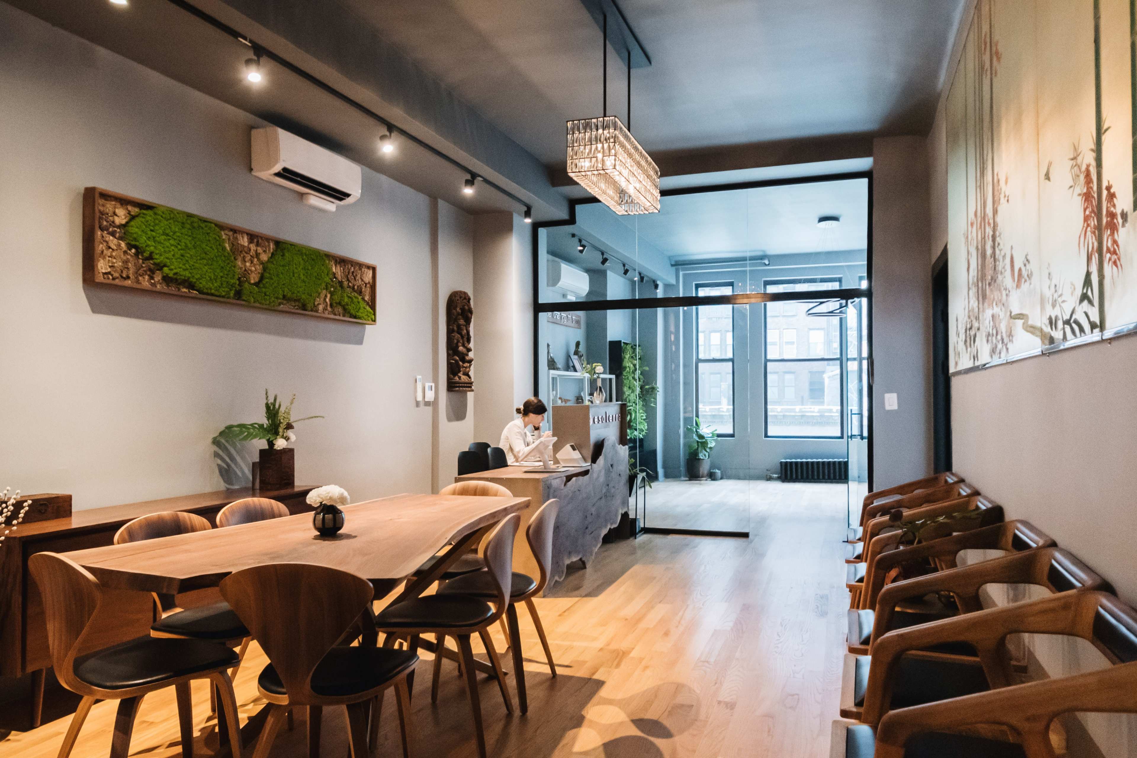 A modern, minimalistic dining area features a large wooden table surrounded by chairs, with a person working at a desk in the background.