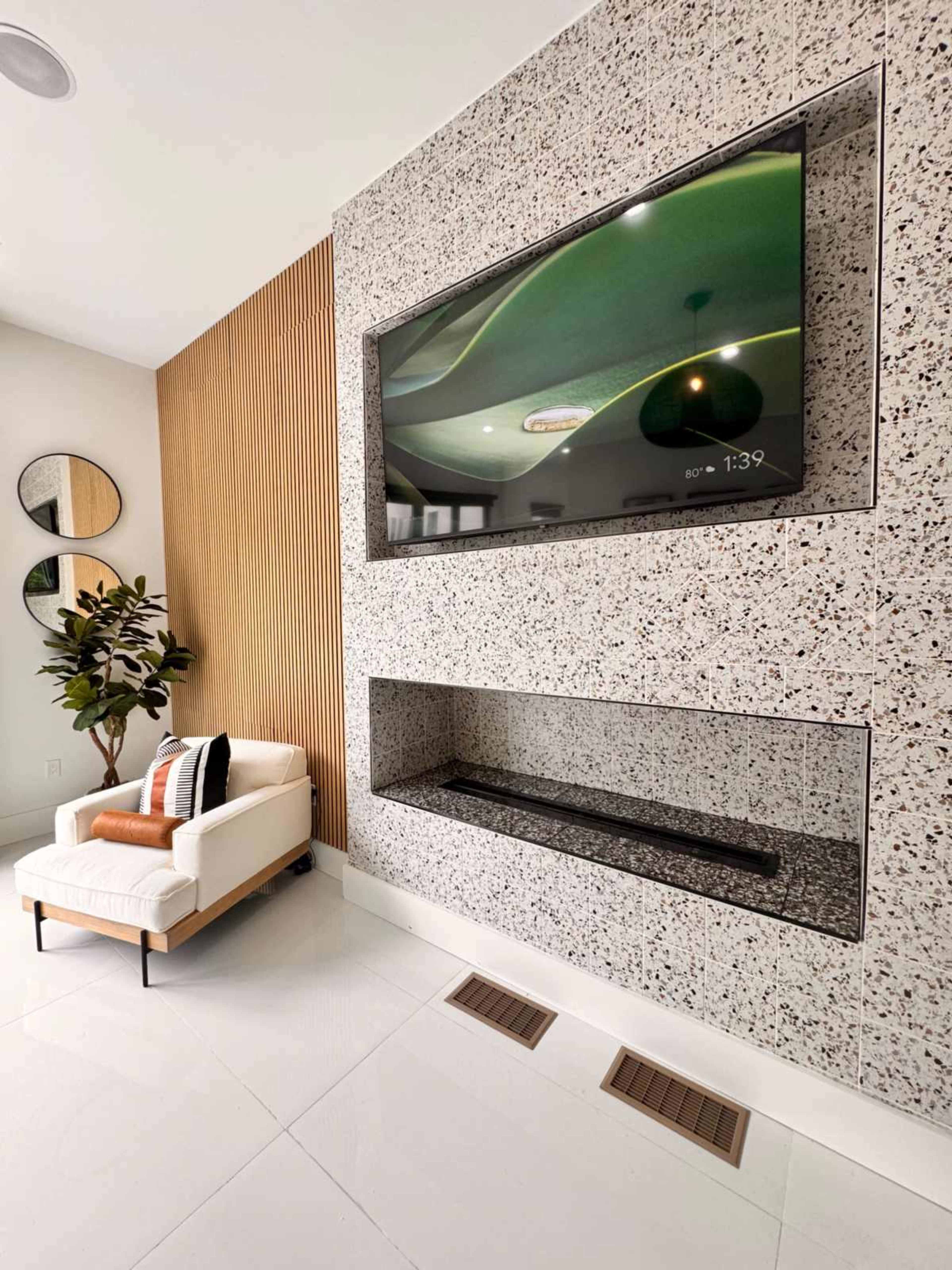 The image shows a modern living room featuring a textured wall with a mounted television above a fireplace and a single armchair nearby.