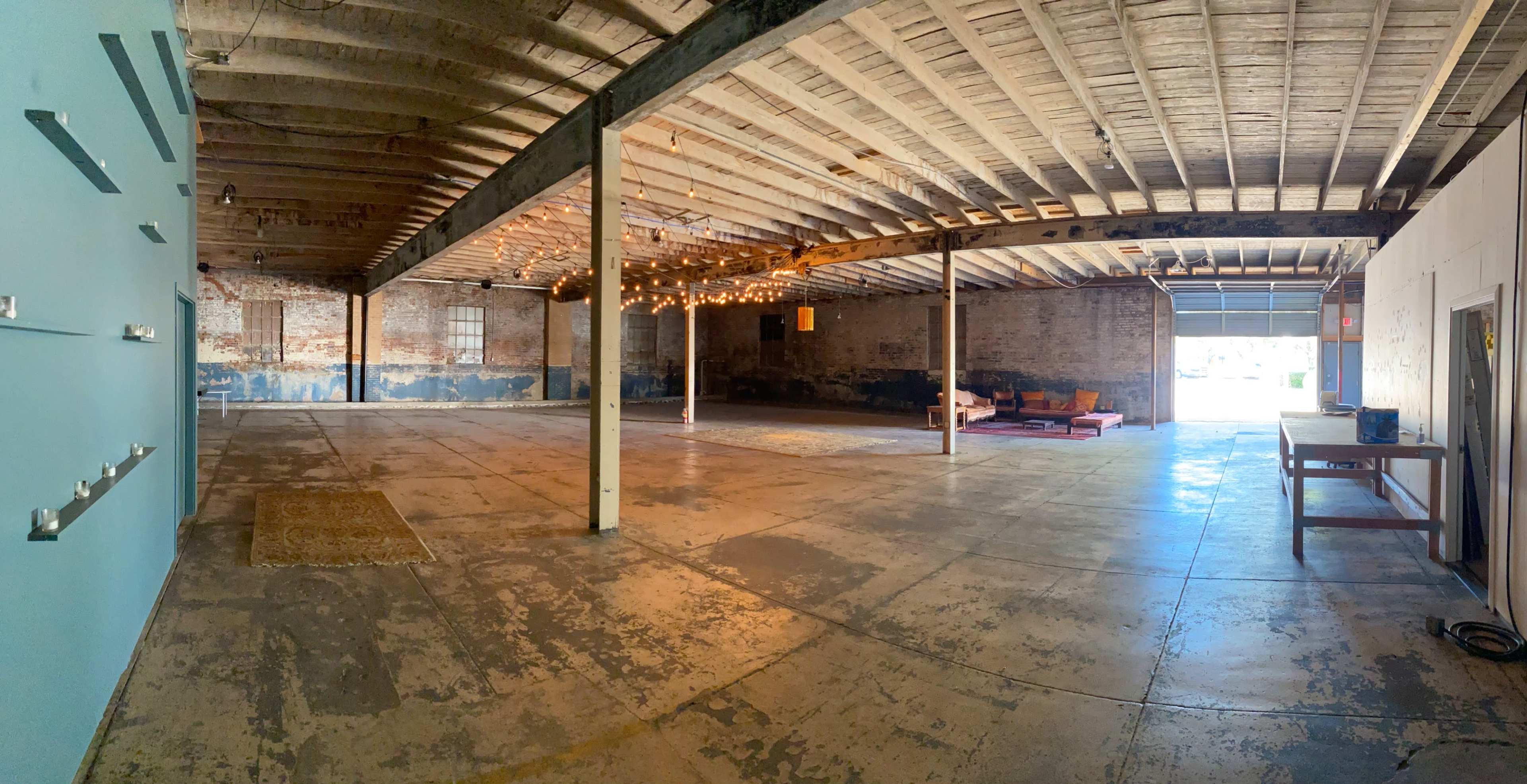 The image shows a spacious, industrial-style room with exposed beams, a concrete floor, and a section featuring soft lighting and a seating area.