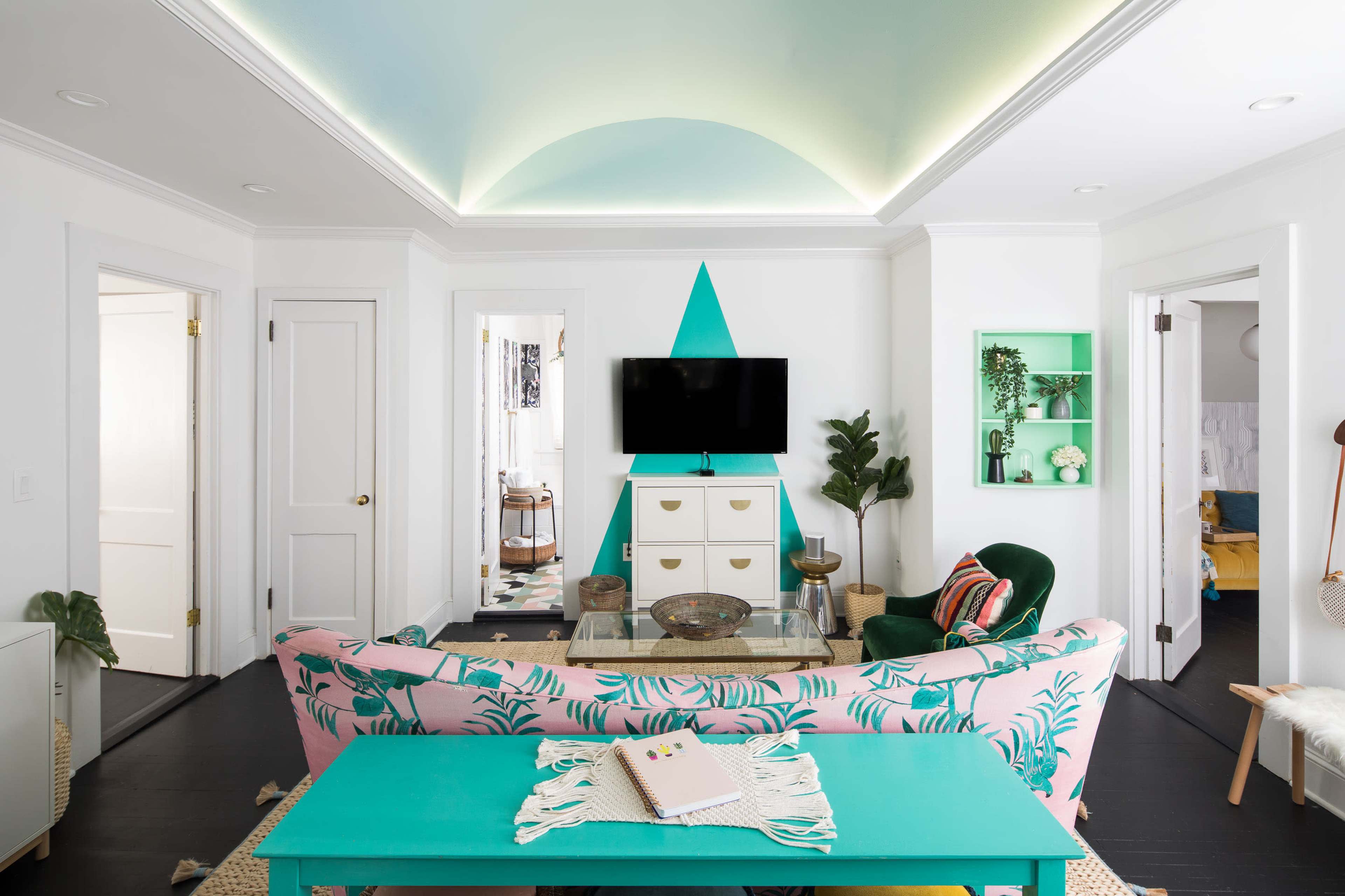 A brightly decorated living room features a pink sofa with tropical patterns, an aqua coffee table, and a mounted television against a geometric accent wall.