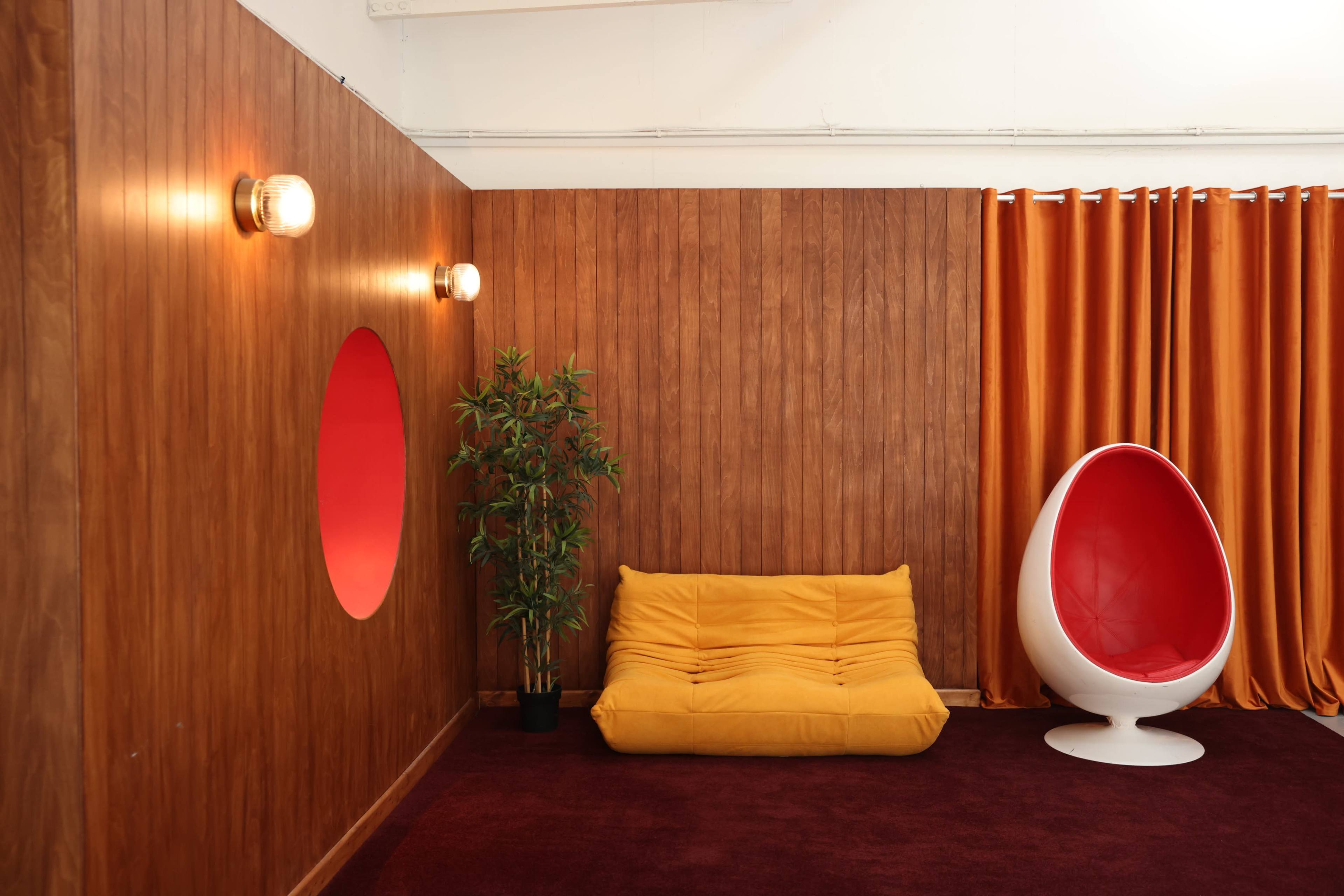 The image shows a room with wooden paneling, a yellow cushioned sofa, a white egg-shaped chair, a potted plant, and orange curtains.