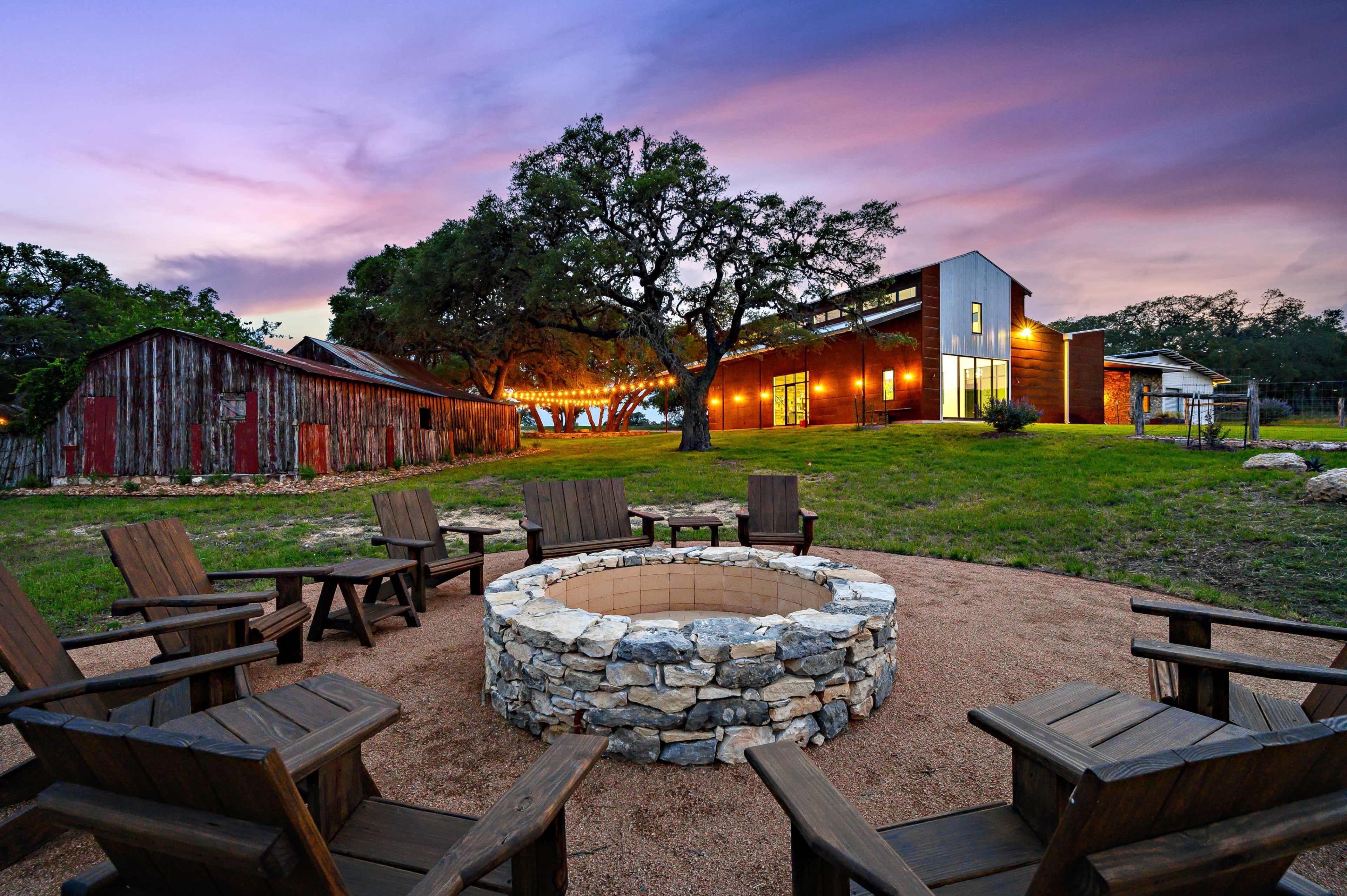 A stone fire pit surrounded by wooden chairs is situated in front of a modern house and rustic barn at sunset.