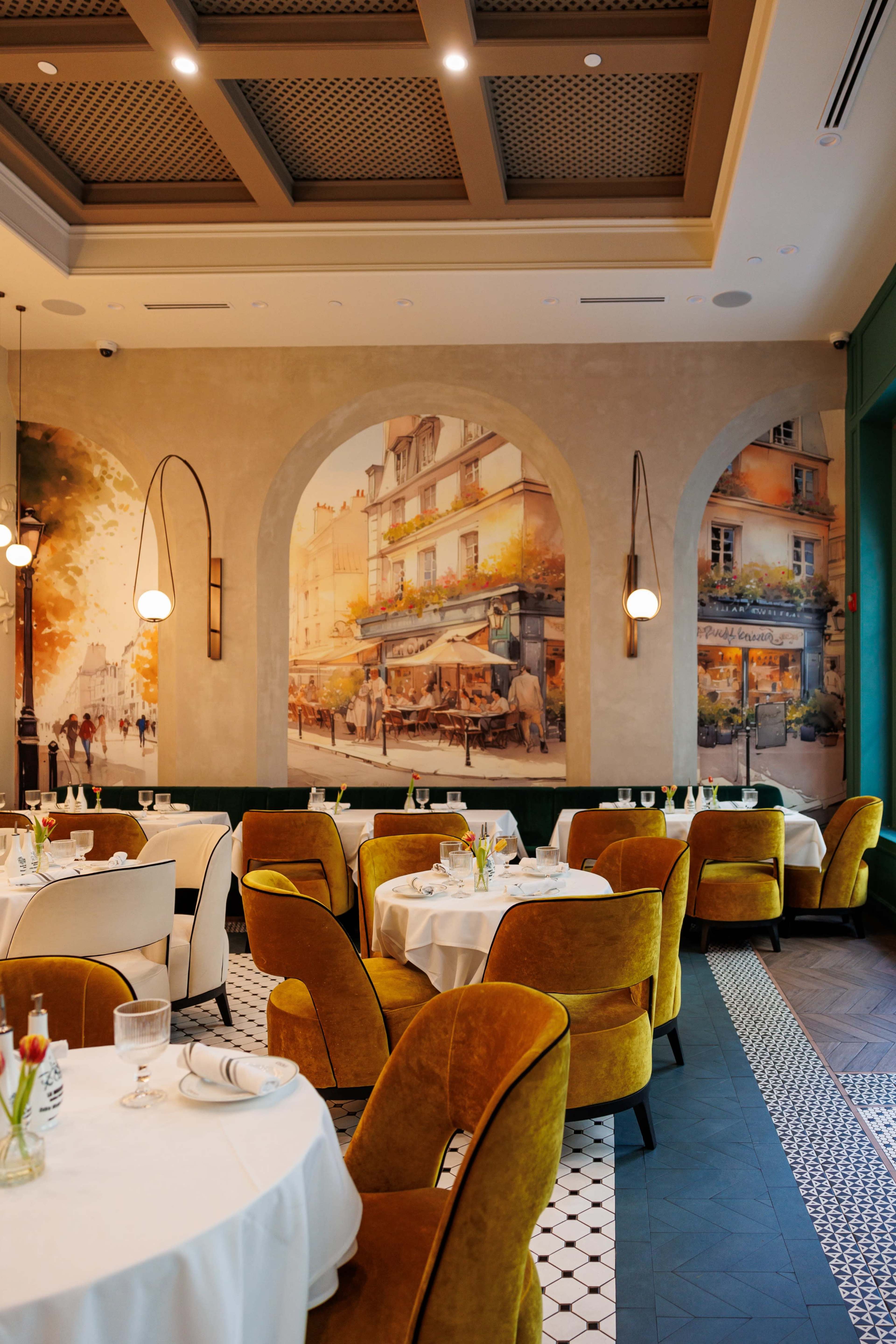 The image shows a restaurant interior featuring elegant tables set with white linens and yellow velvet chairs, complemented by a decorative mural of a street scene on the wall.
