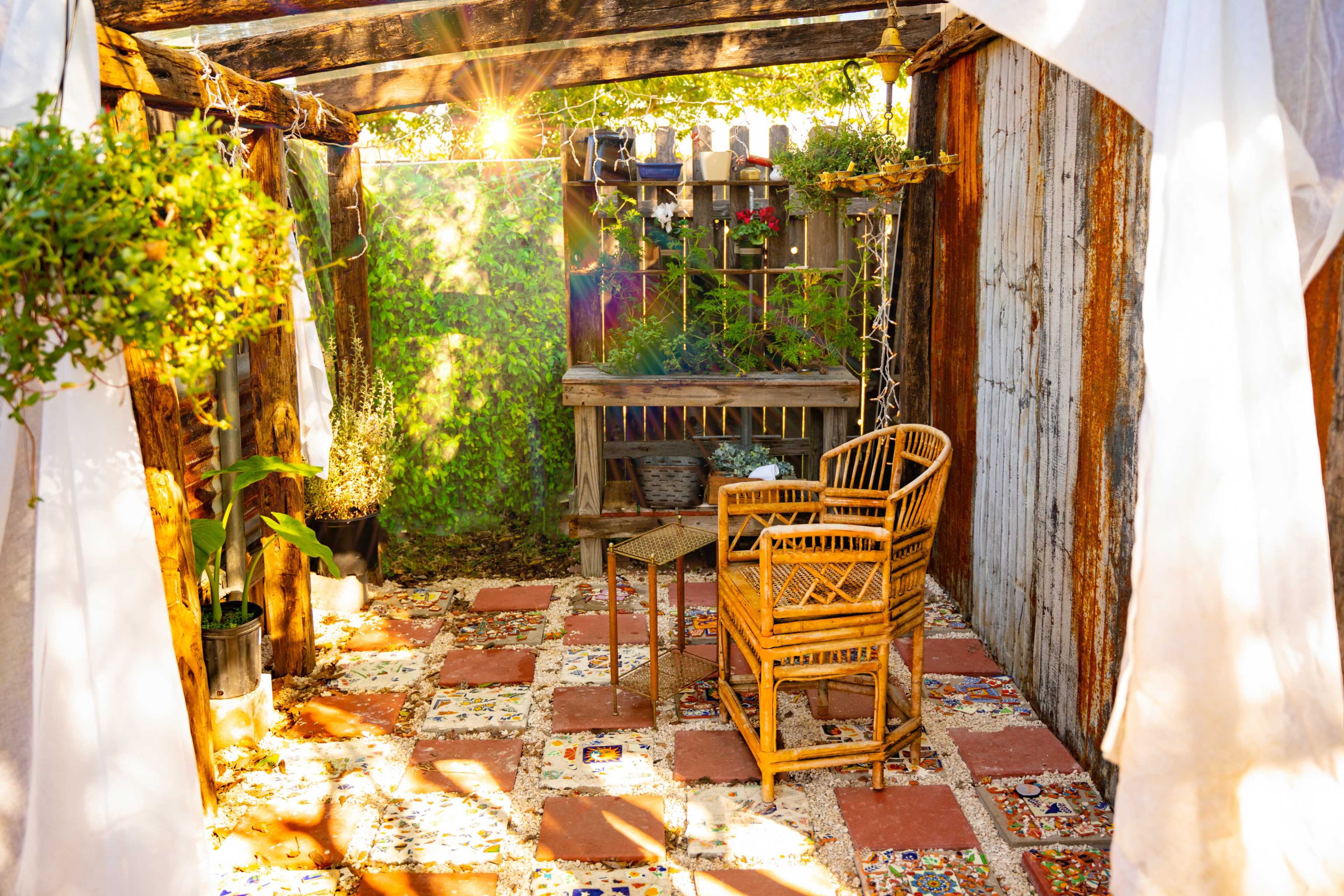 A cozy outdoor seating area features a rattan chair and small table surrounded by greenery and colorful tiled flooring.