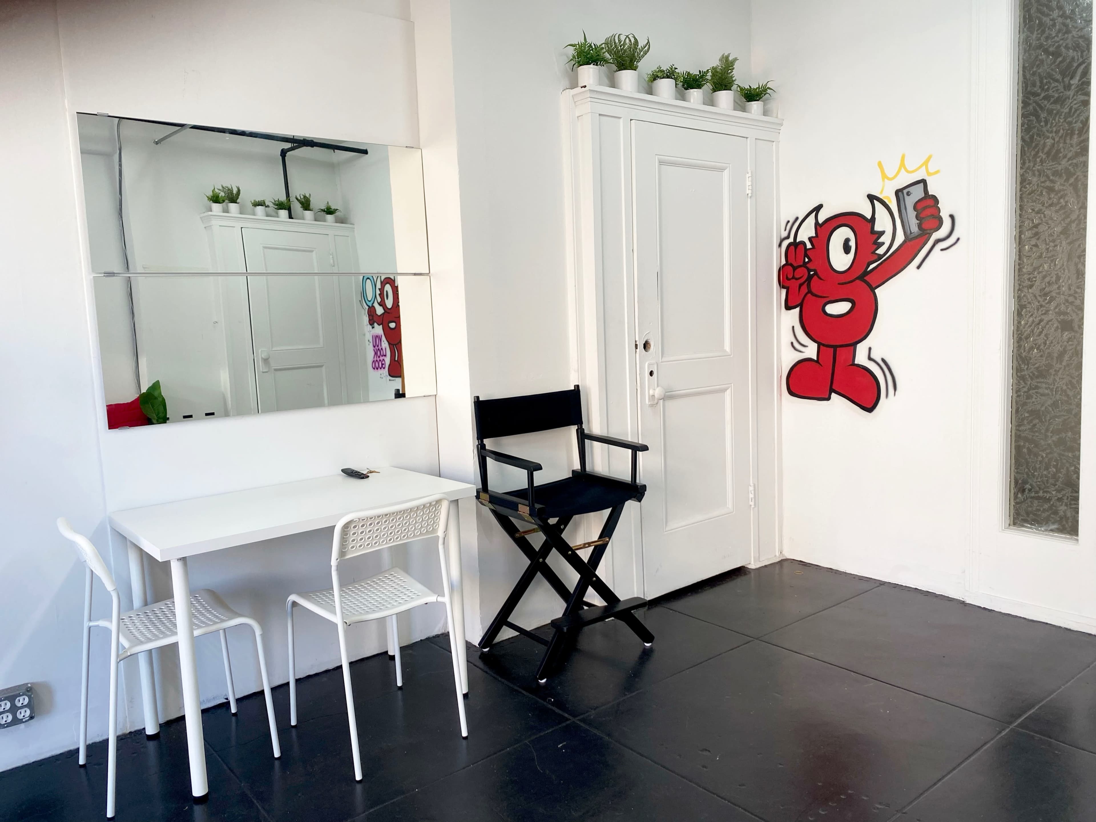 A minimalist room with a white table and two chairs, a large mirror on the left wall, and a door adorned with a red cartoon figure holding a phone.