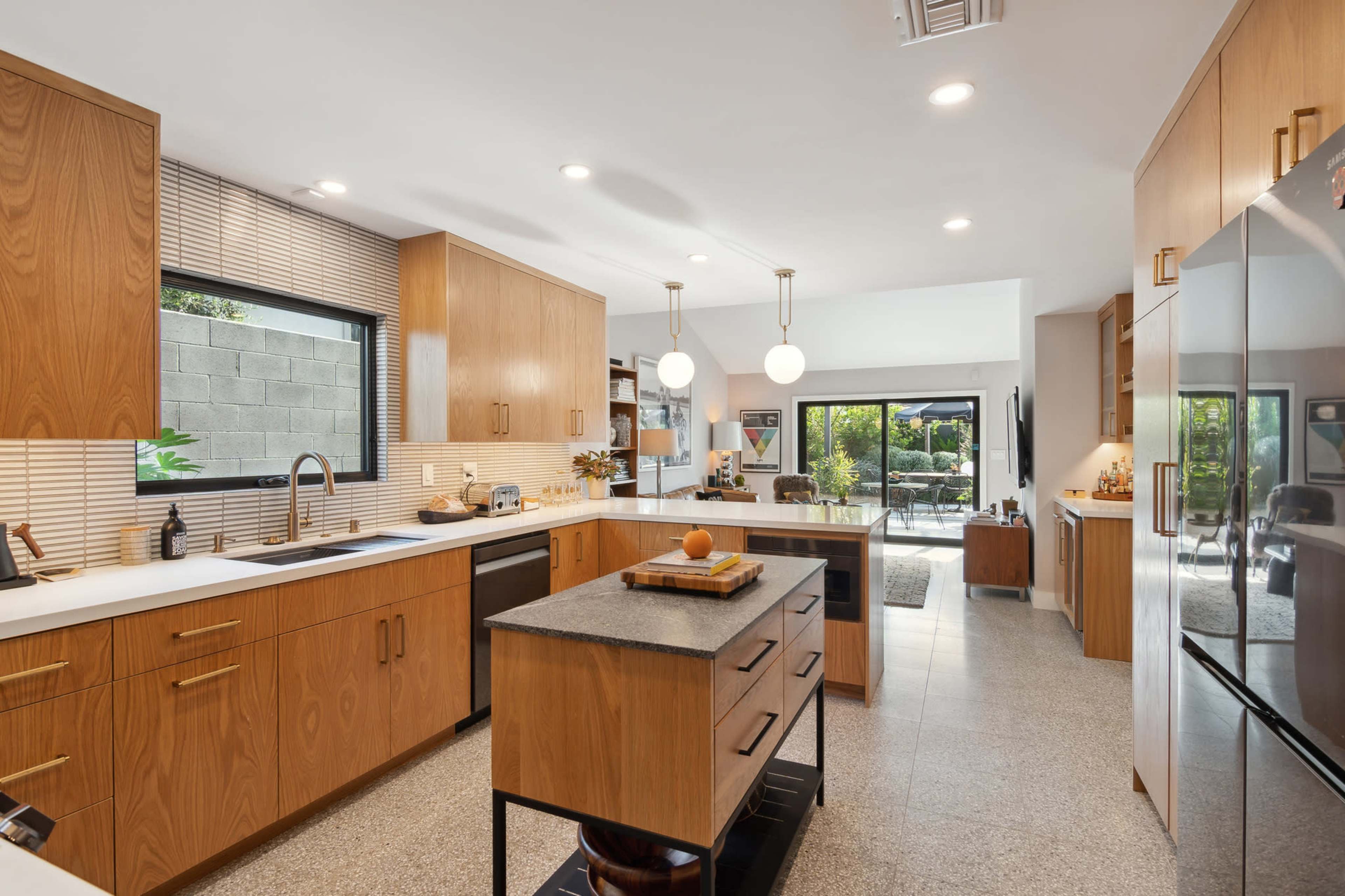 A modern kitchen features light wood cabinetry, a central island with a dark countertop, and large windows that overlook a patio area.