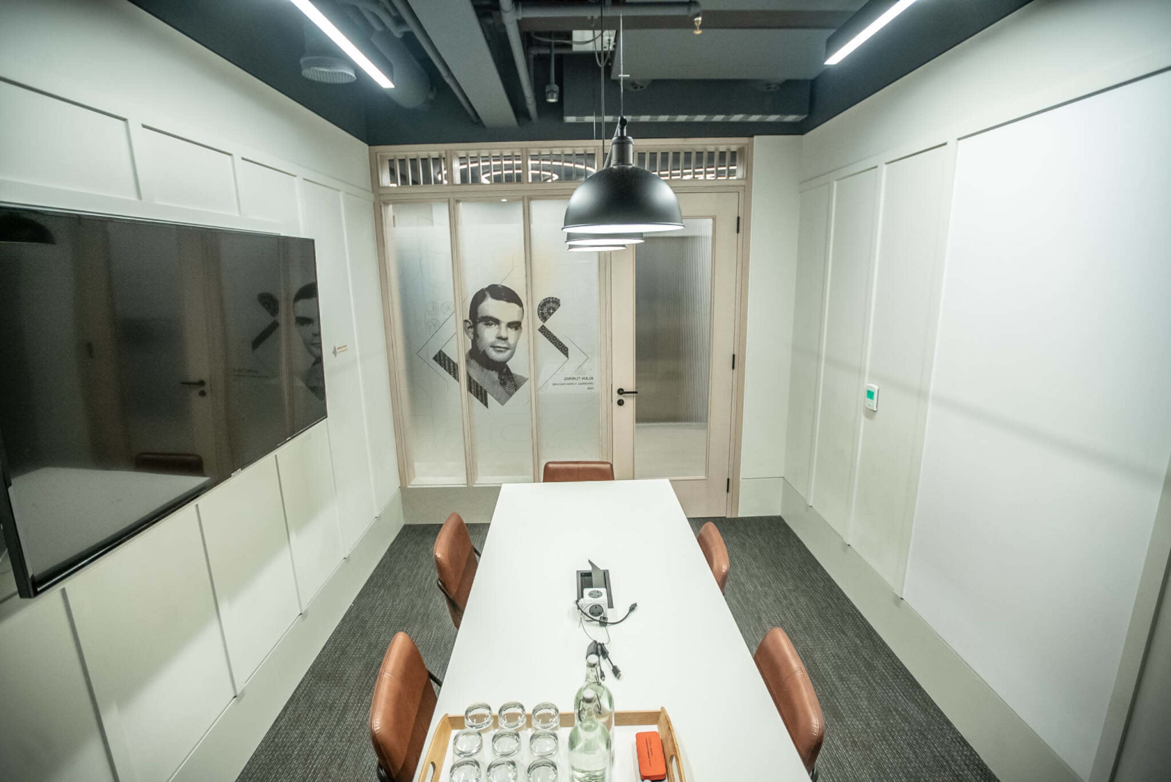 The image shows a modern conference room featuring a central table with chairs, a television on one wall, and a glass partition displaying a printed design.