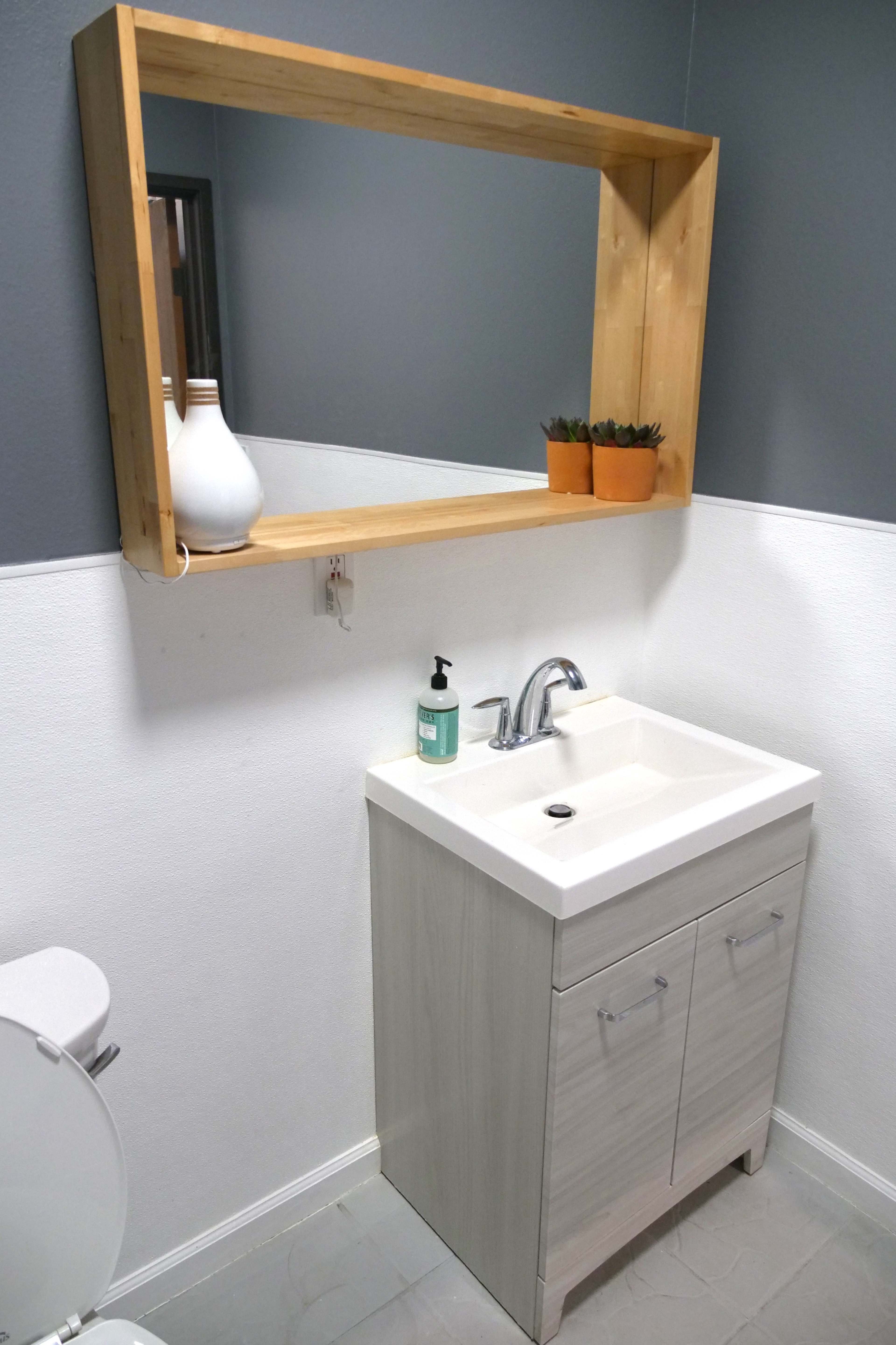 A modern bathroom featuring a wooden-framed mirror above a light-colored vanity with a white sink and two storage drawers, beside a toilet.