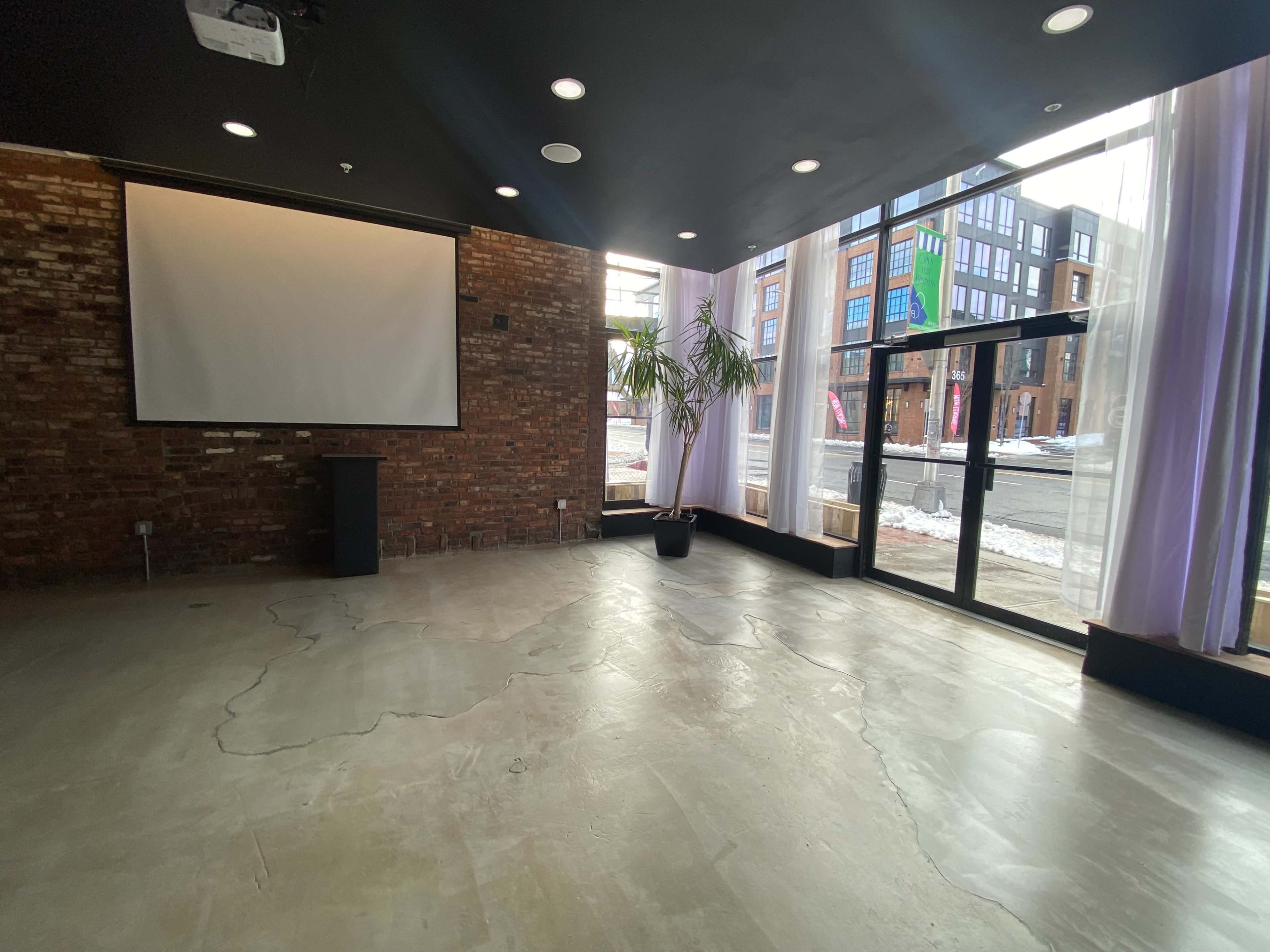 The image shows a spacious room with exposed brick walls, a projector screen, and large windows facing a street.