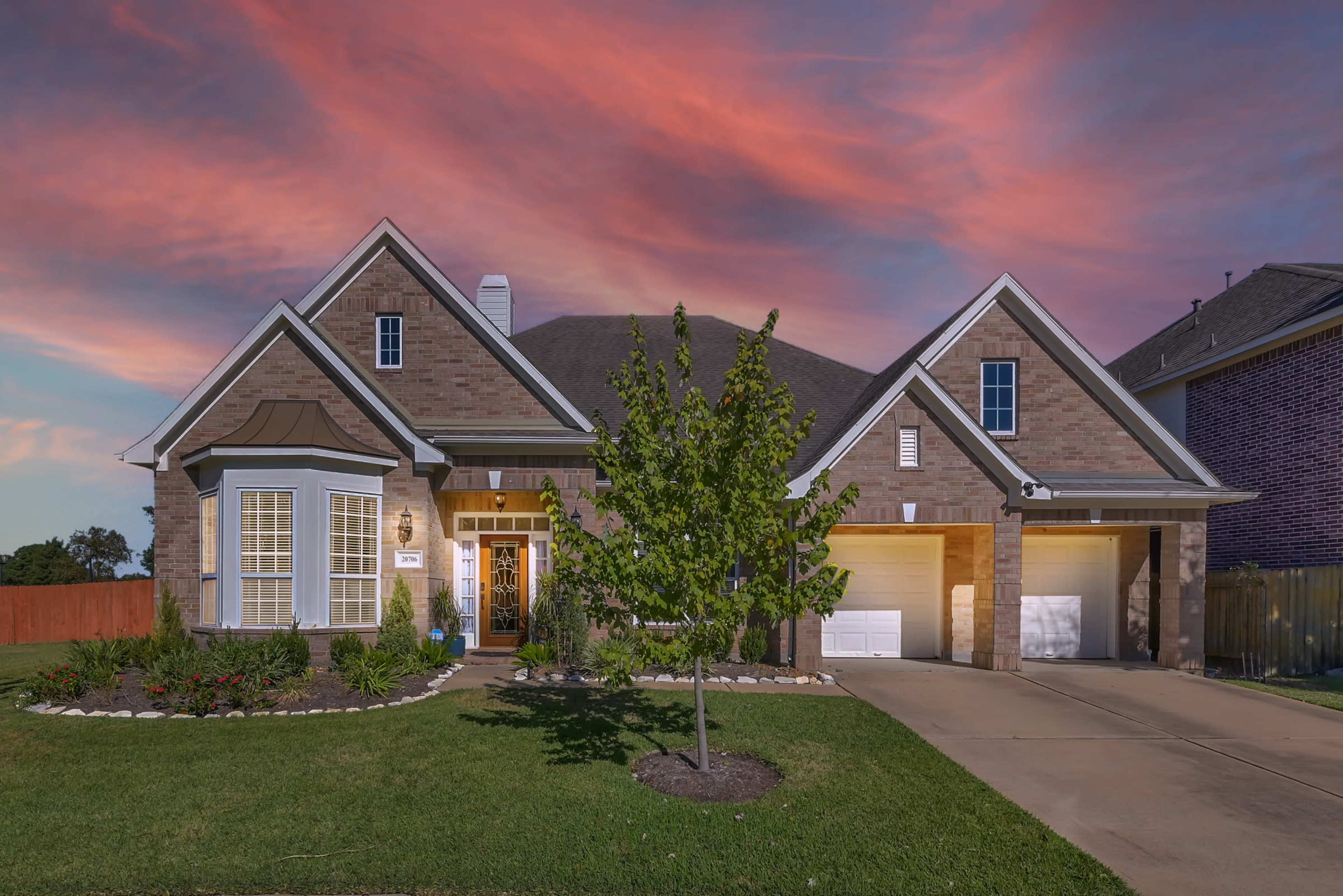 A single-story brick house with a manicured lawn and a driveway leading to two garages is illuminated under a colorful sunset sky.