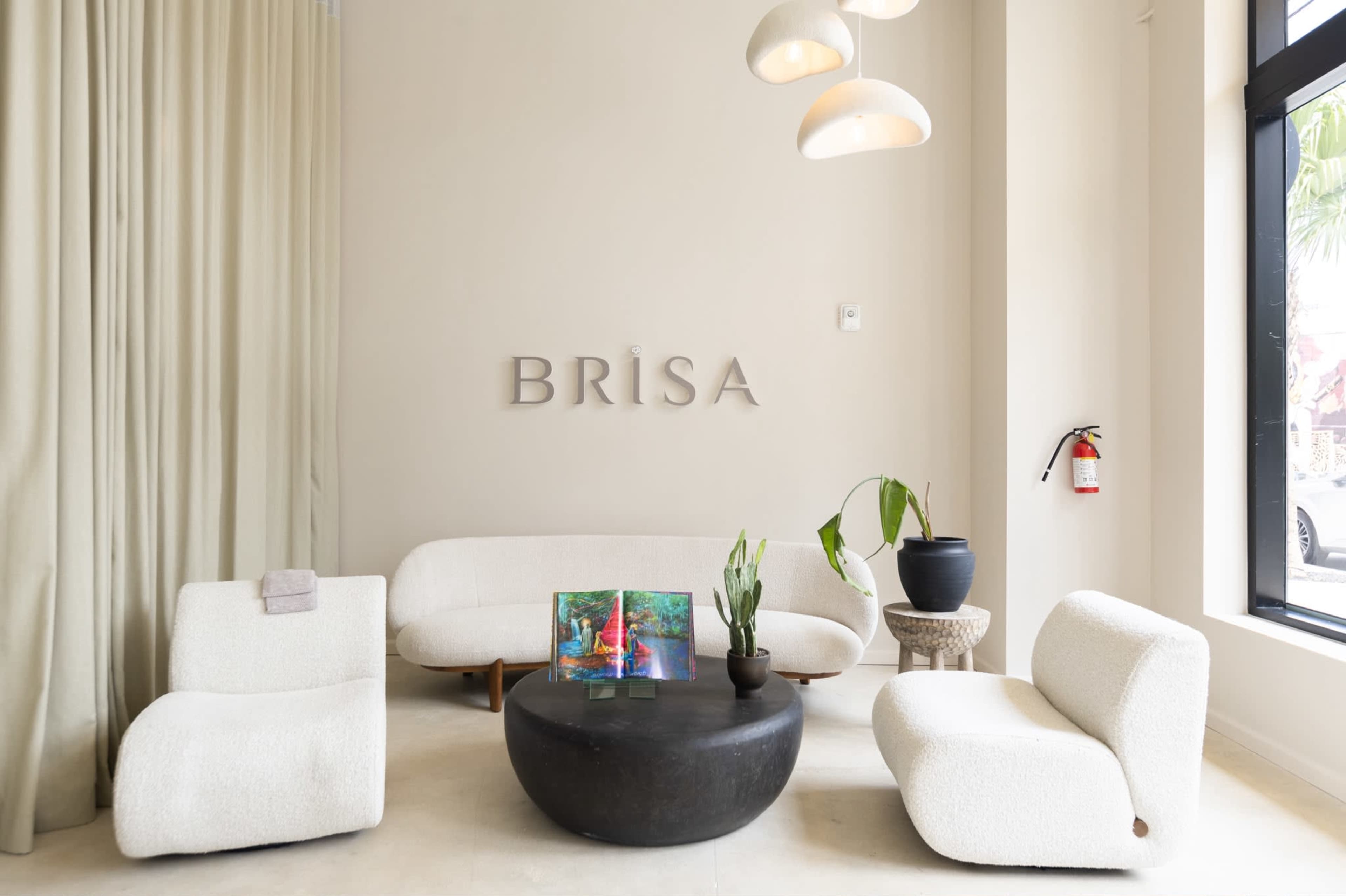 The image shows a minimalist reception area with two curved white sofas, a black round coffee table, and a small plant next to a window, featuring the name "BRÏSA" on the wall.