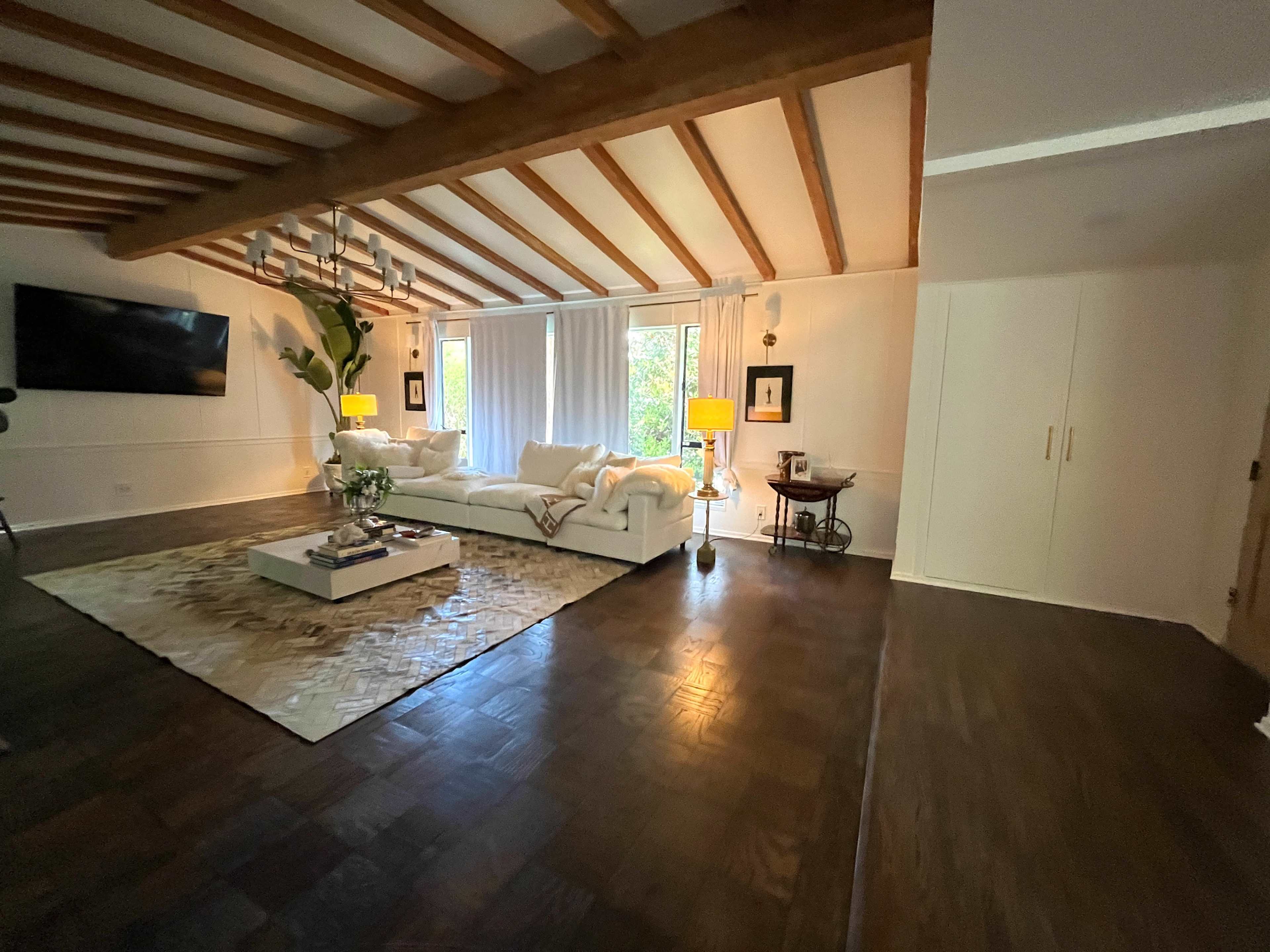 A spacious living room features a white sectional sofa, large windows with sheer curtains, and wooden ceiling beams.