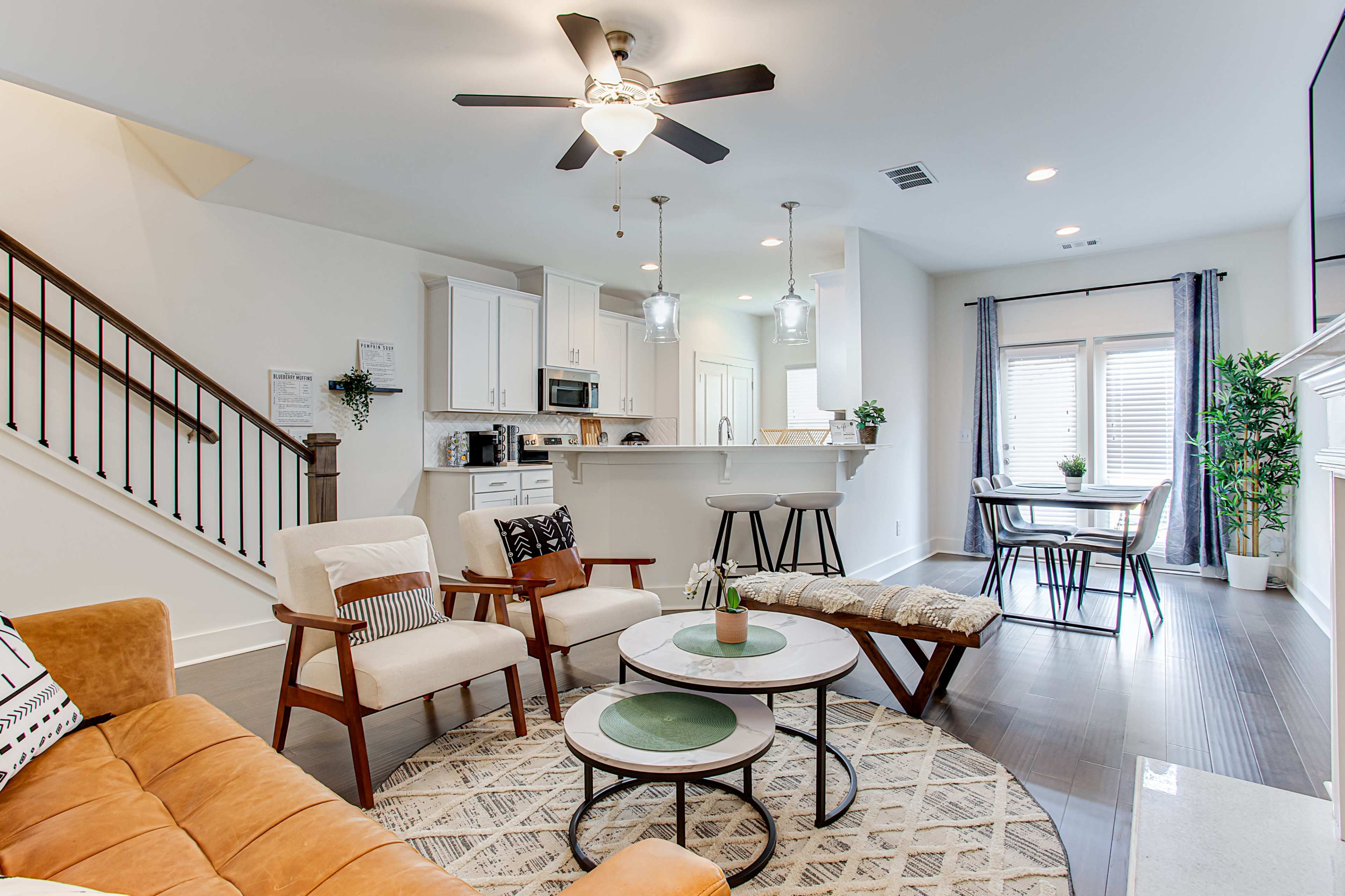 A modern living room features a tan couch, white chairs, a round coffee table, and a dining area in the background with a table and chairs.
