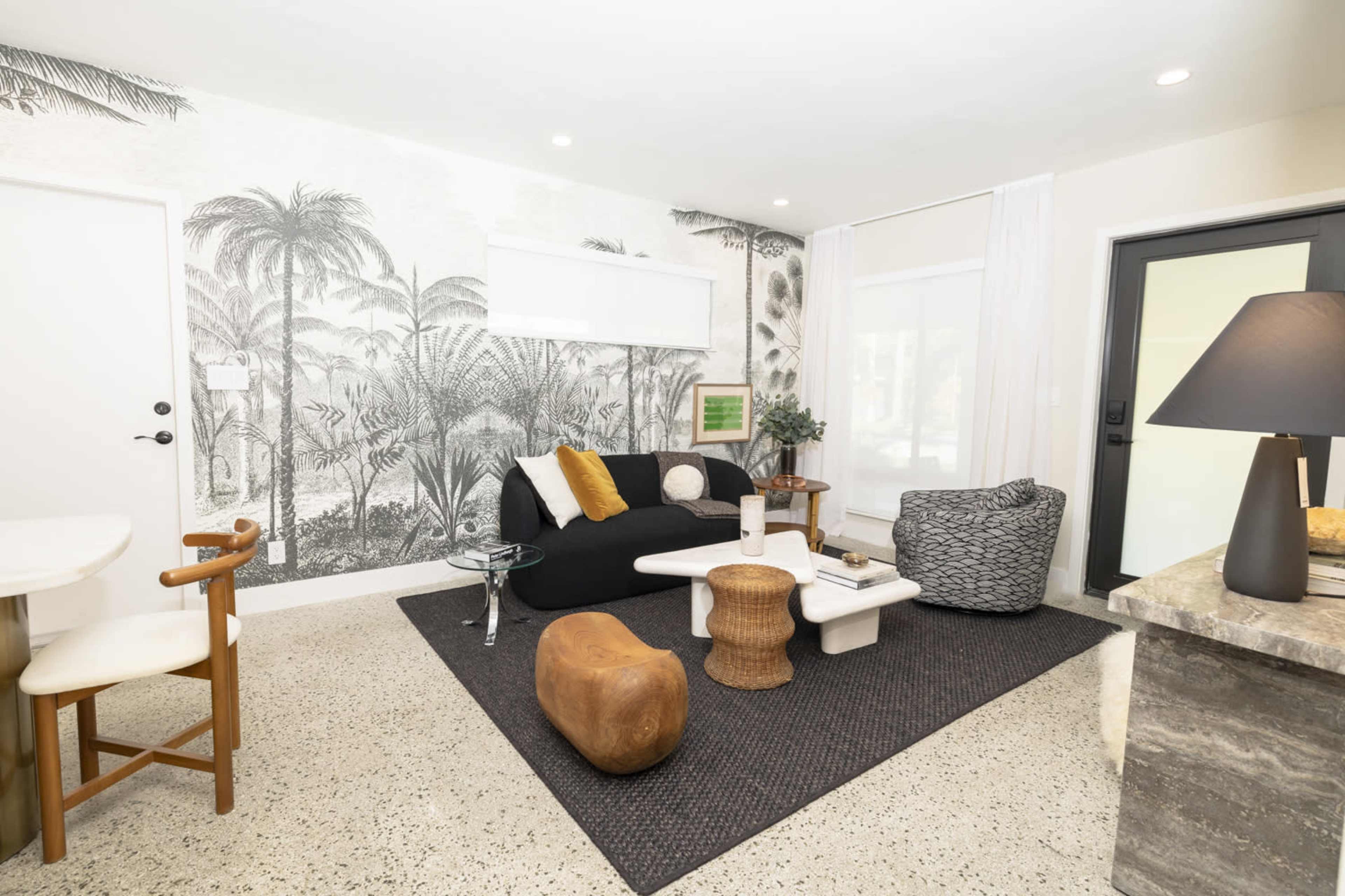 A modern living room features a black sofa, textured armchair, a white coffee table, and a large tropical mural on the wall.