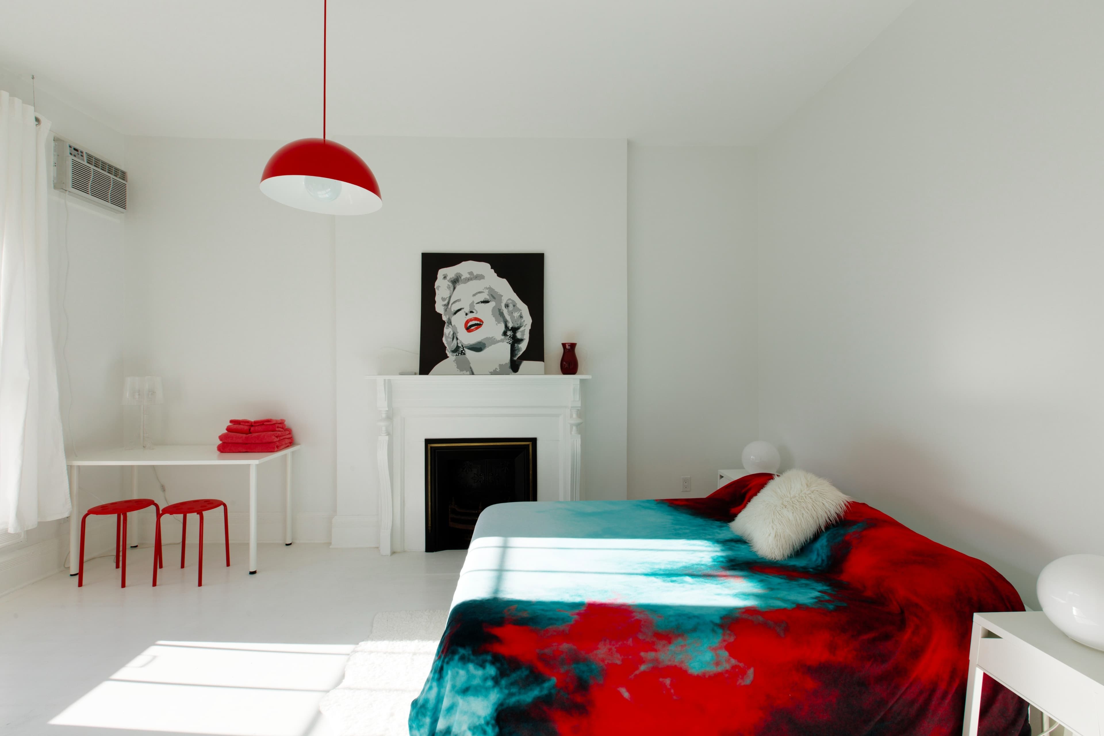 A minimalist bedroom features a bed with a colorful blanket, a white fireplace, a red pendant light, and a black-and-white artwork of a woman on the wall.