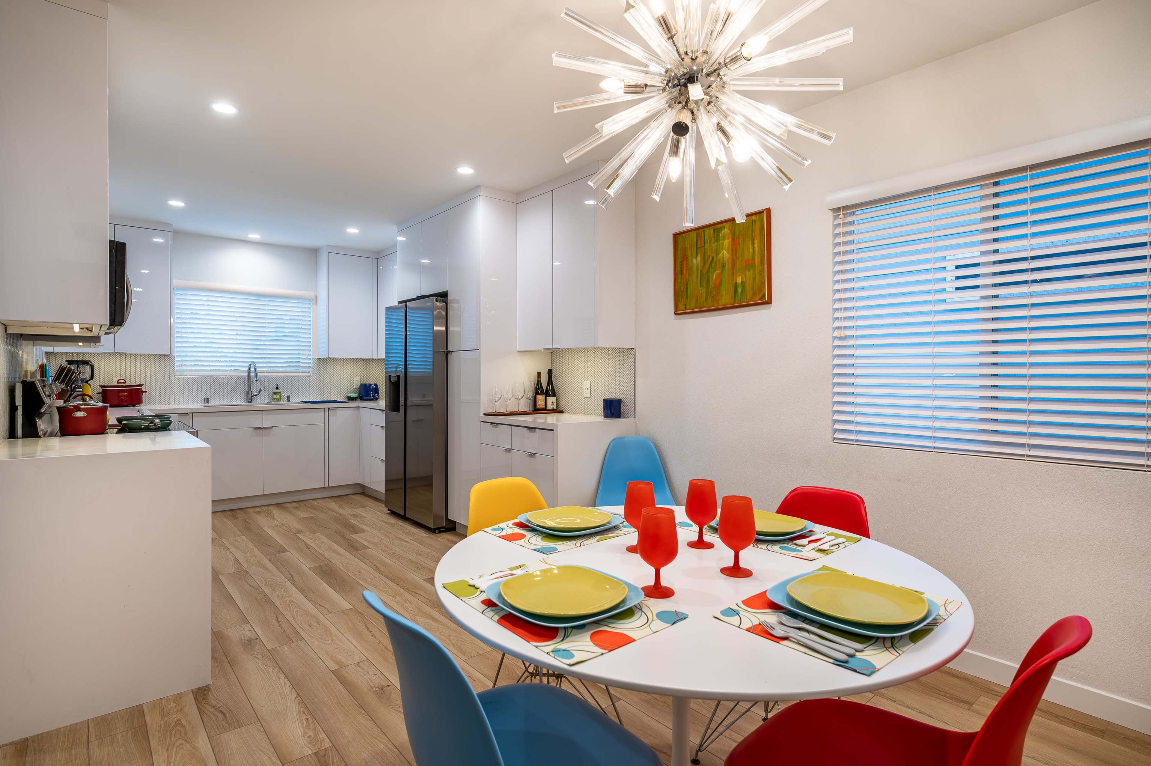 A modern kitchen features a circular dining table set with colorful plates and glasses, surrounded by vibrant chairs, with sleek cabinetry and an organized countertop in the background.