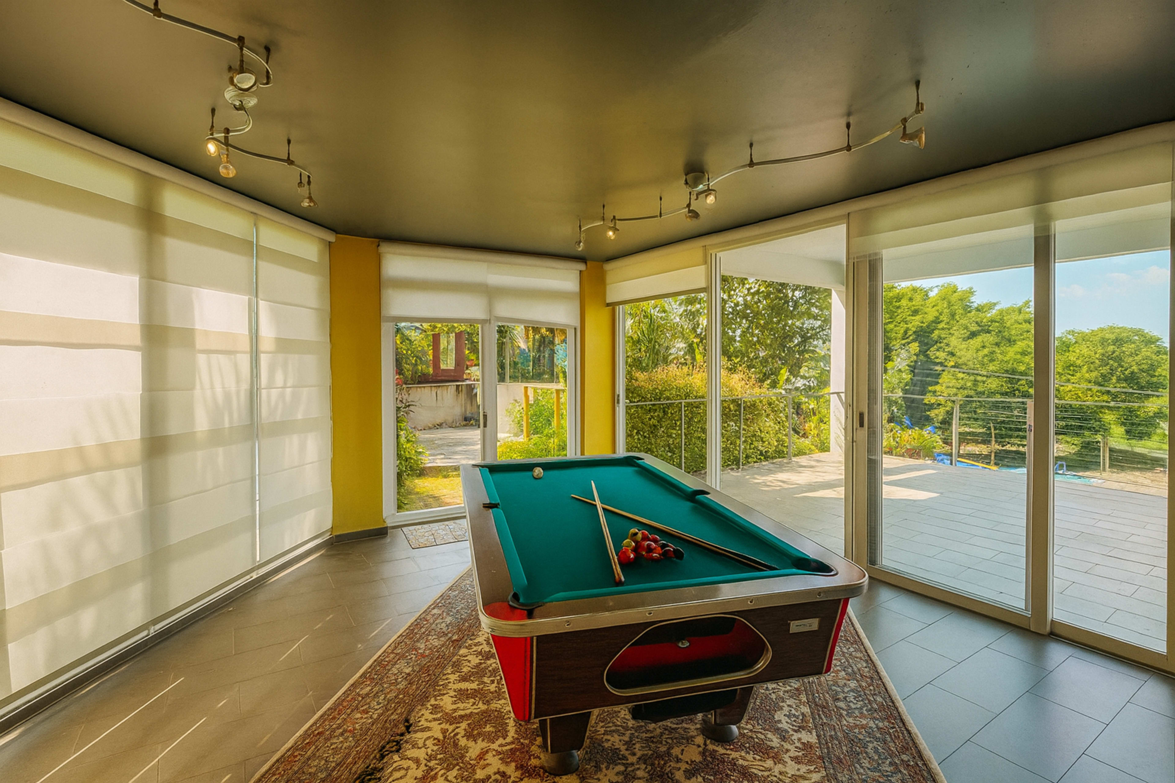 The image shows a brightly lit room with large windows, a green felt pool table in the center, and a sliding glass door leading to an outdoor area.