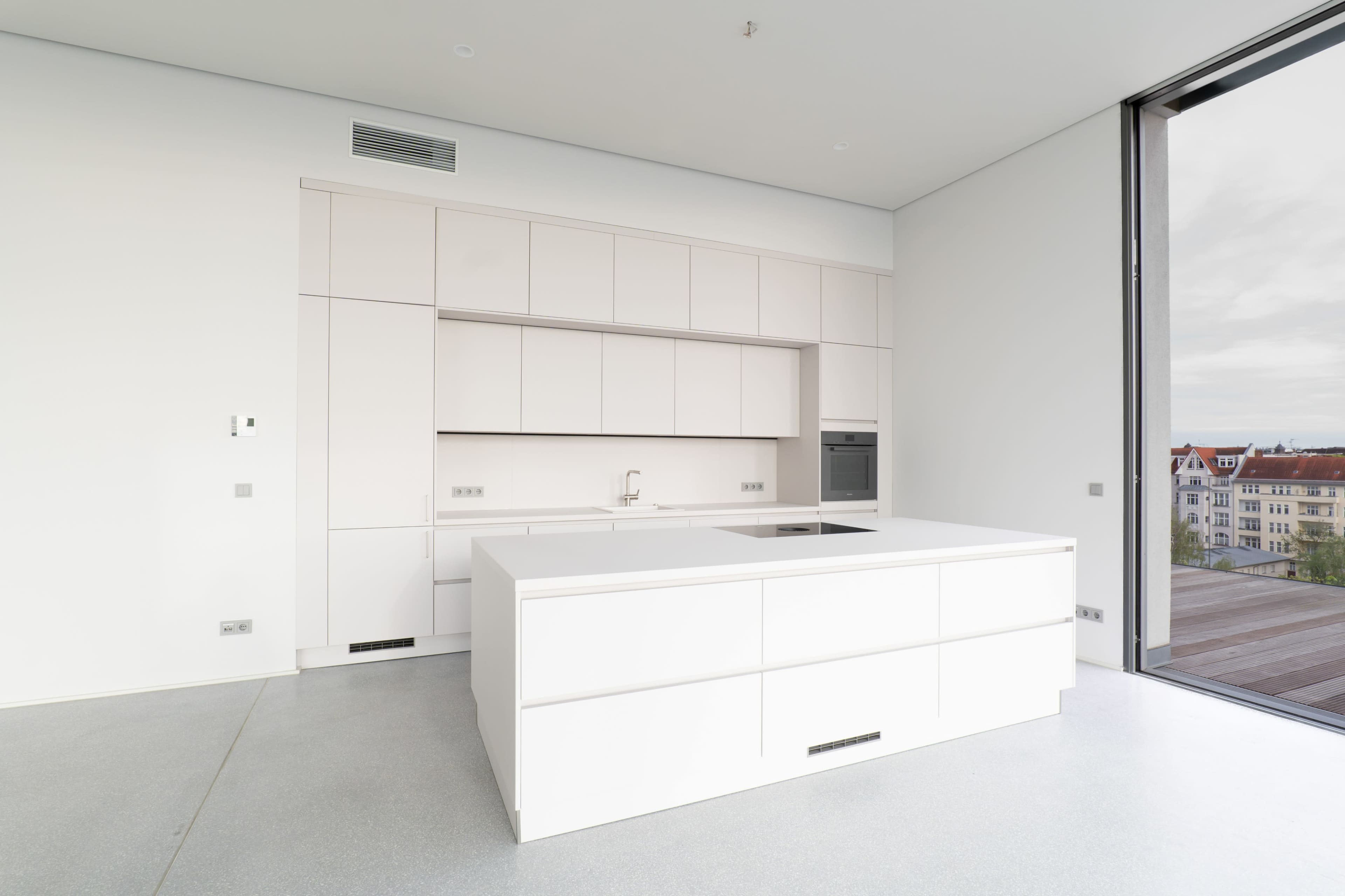 The image shows a modern, minimalist kitchen with a large white island and cabinetry against a plain wall and large windows.