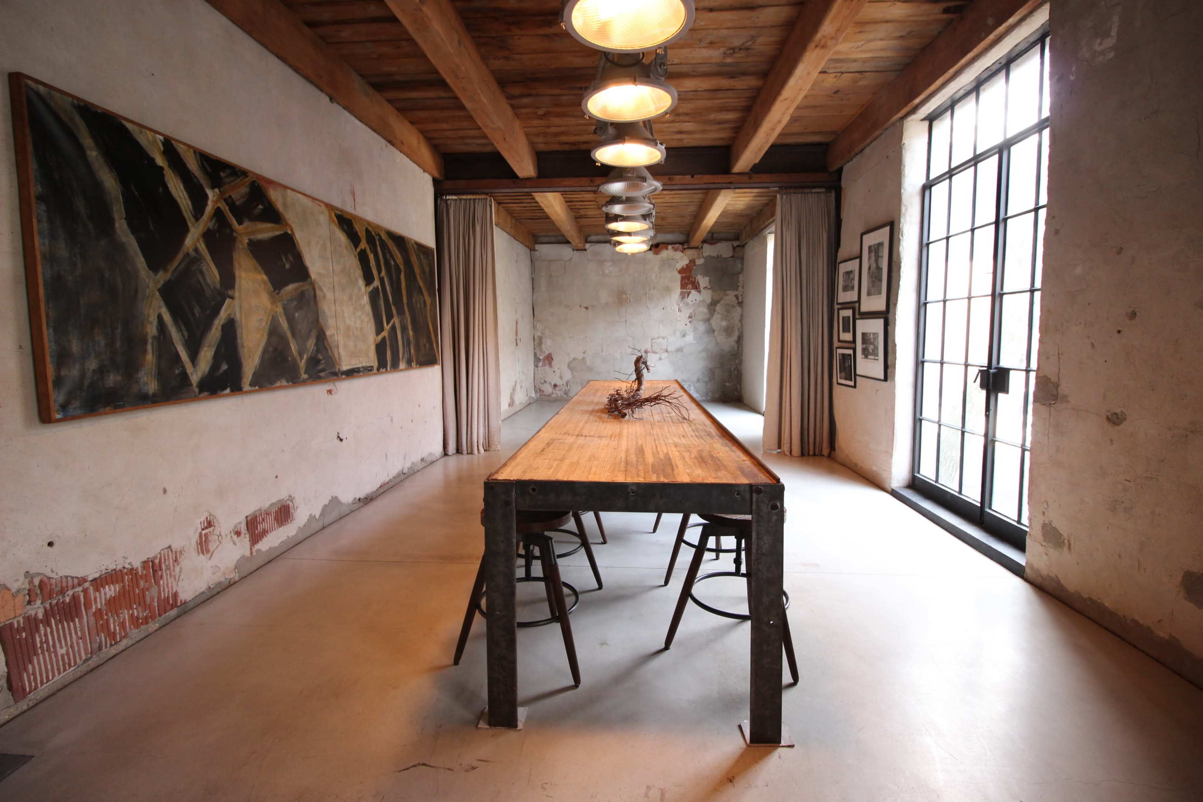 A spacious dining area with a long wooden table, metal chairs, and large windows letting in natural light, framed by minimalistic decor and exposed brick walls.