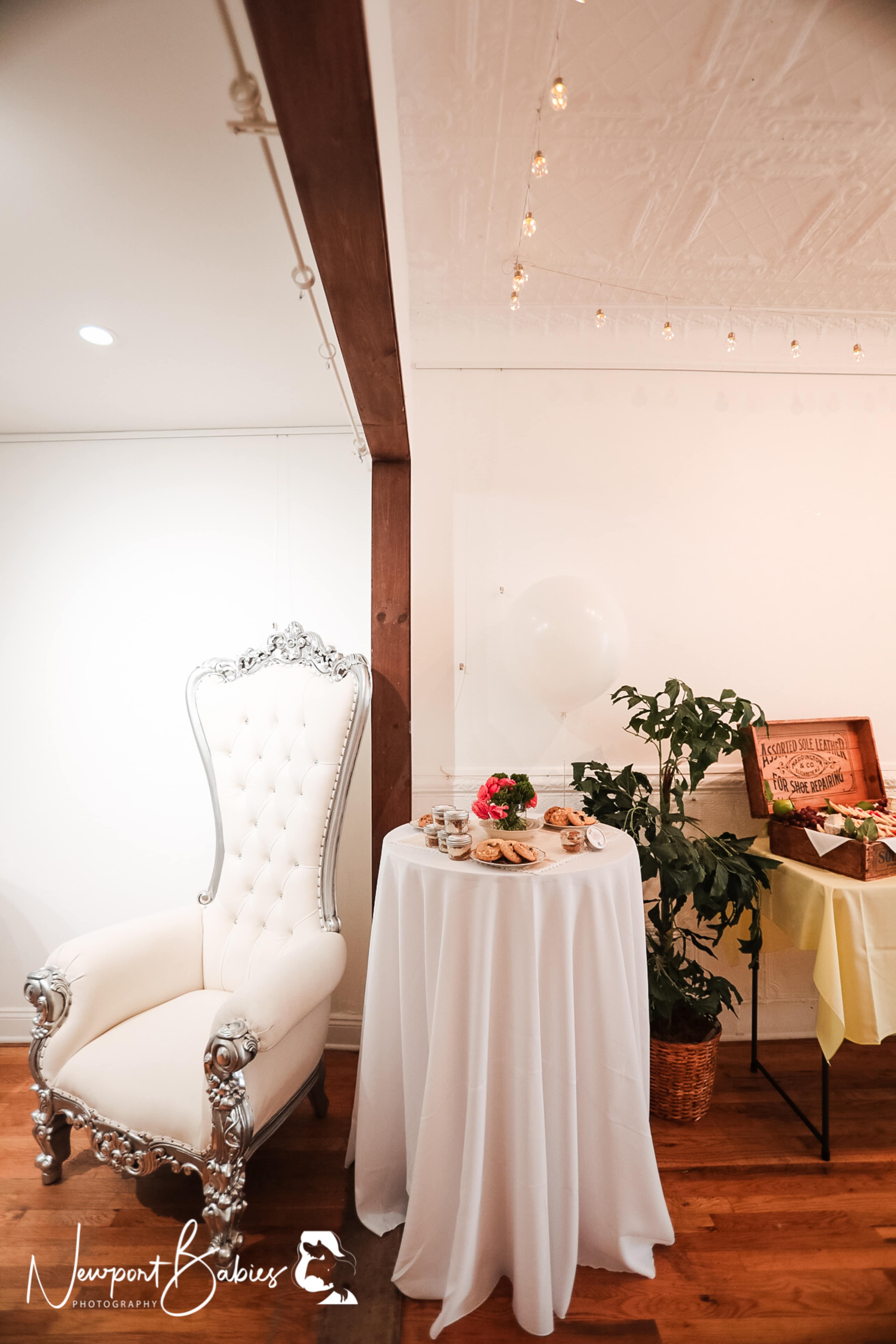A white, ornate chair next to a small round table covered with a white tablecloth displaying an assortment of snacks, alongside a potted plant in a room with wooden flooring and exposed beams.