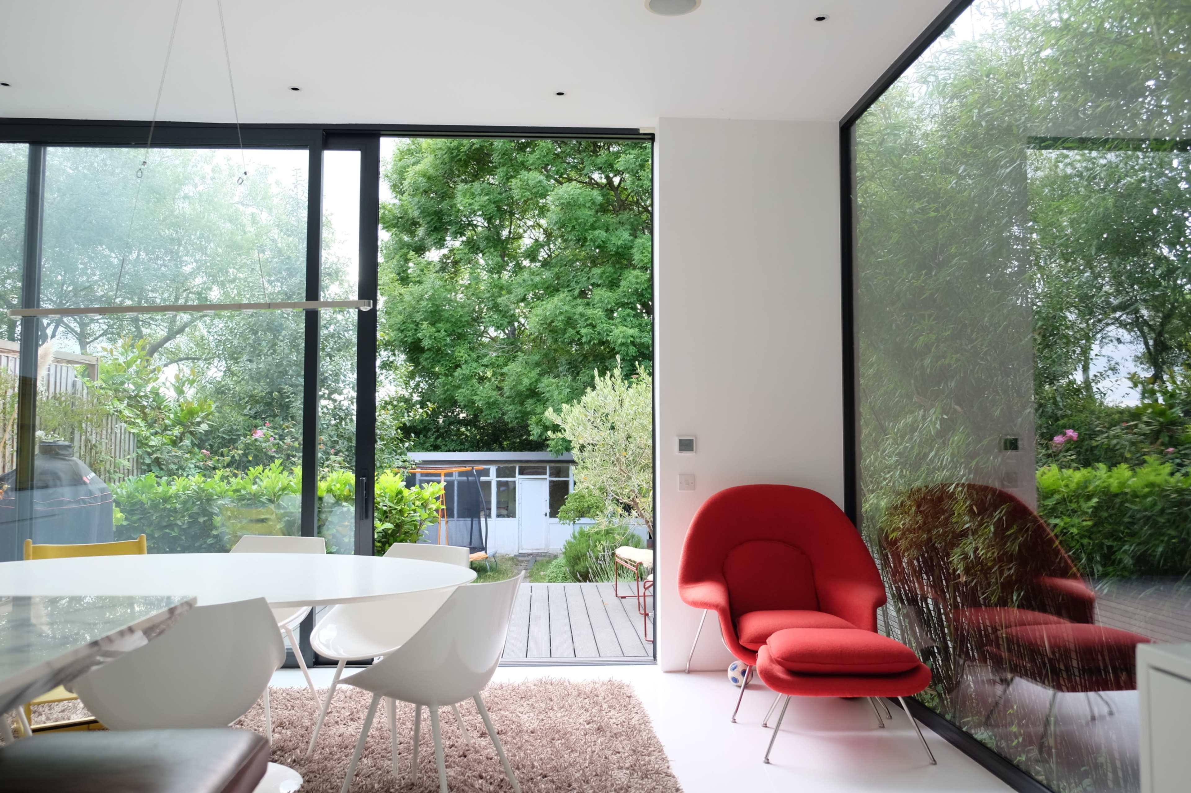 A bright, modern dining area features a round table surrounded by white chairs and a red accent chair, with large windows showcasing a lush green garden outside.