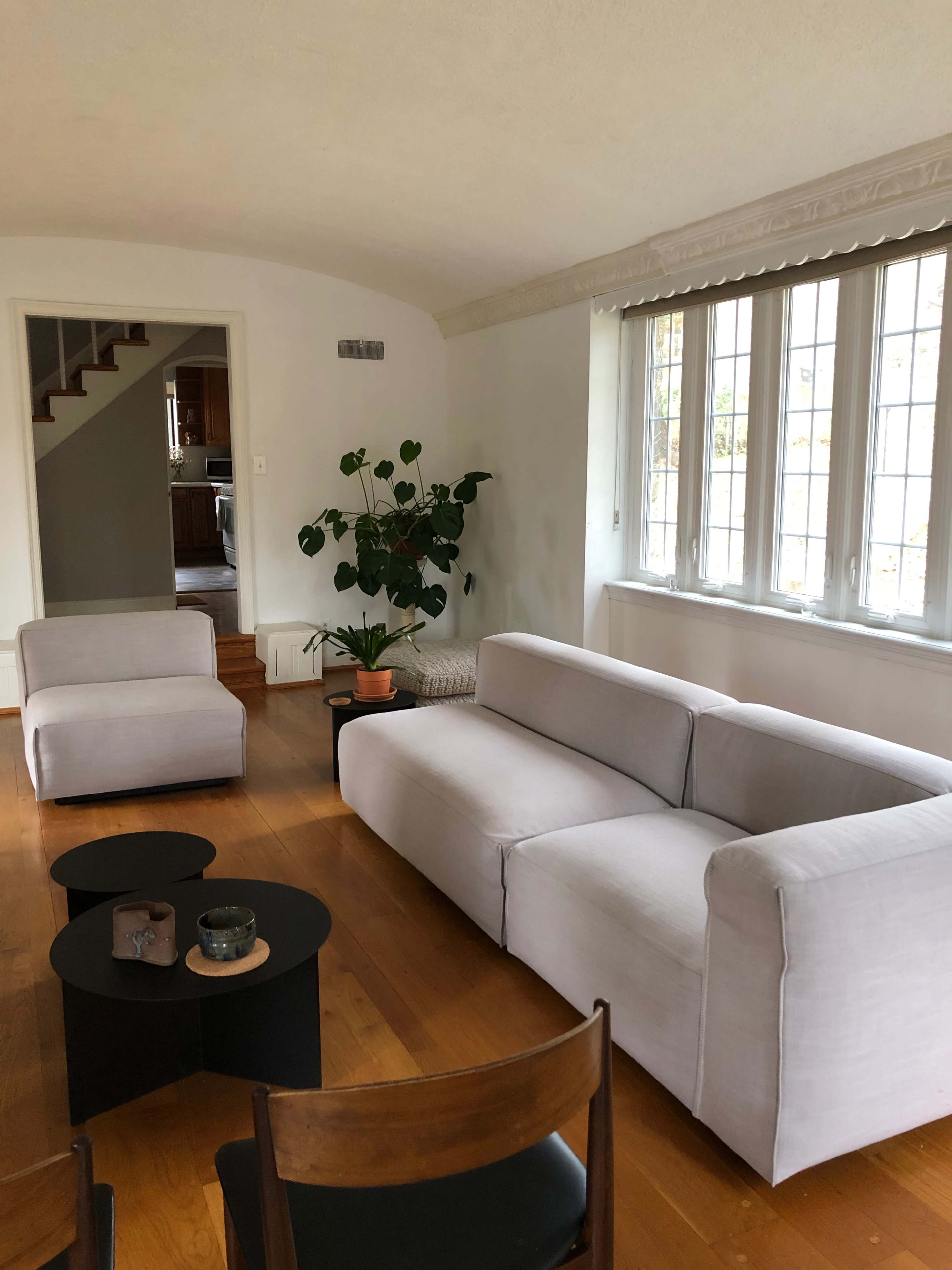A modern living room with a light-colored sofa, a small black table, and two chairs, alongside a potted plant near a window.