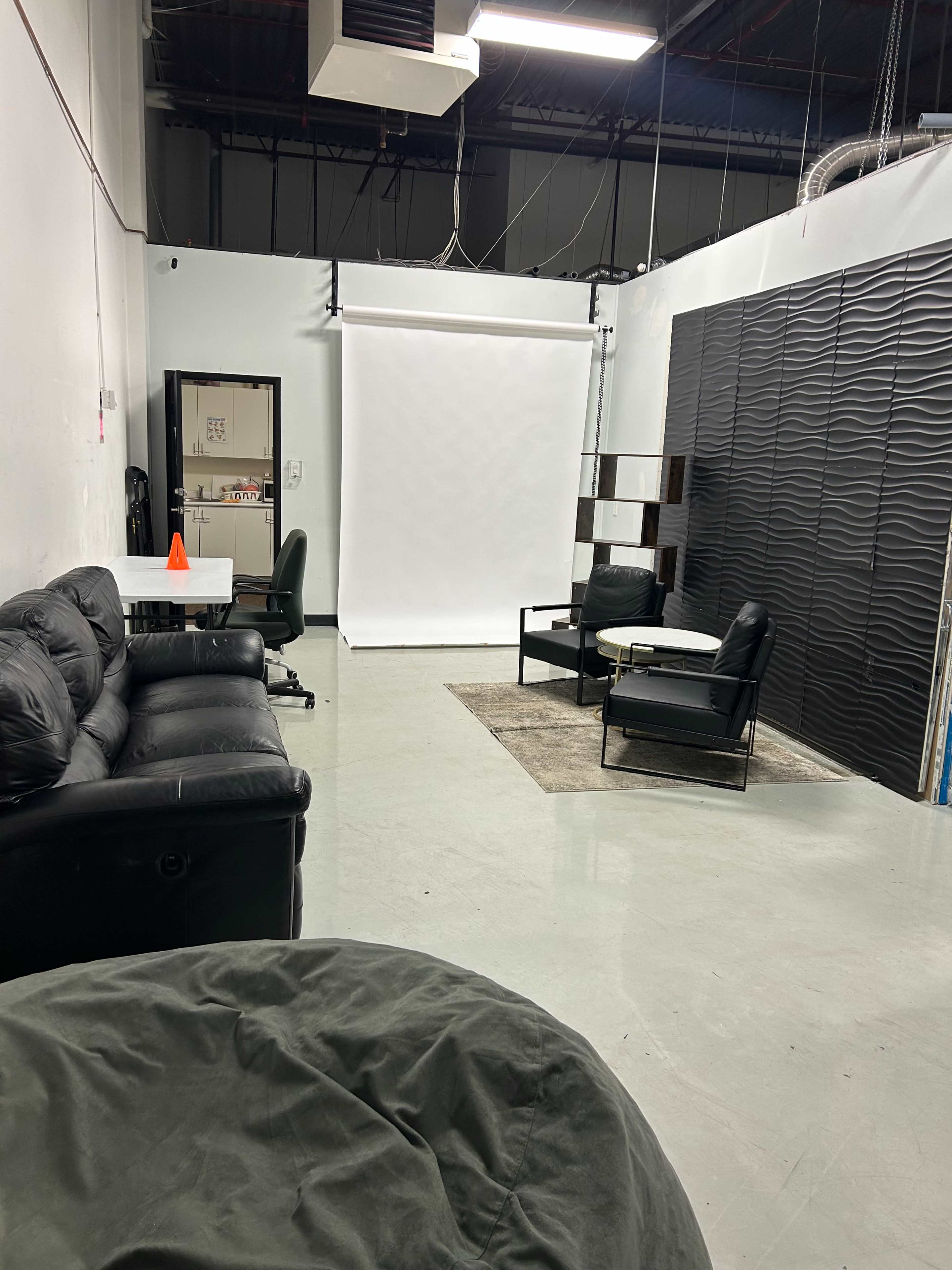 The image shows a spacious, modern room with black sofas, a white backdrop, and a clearly defined seating area.