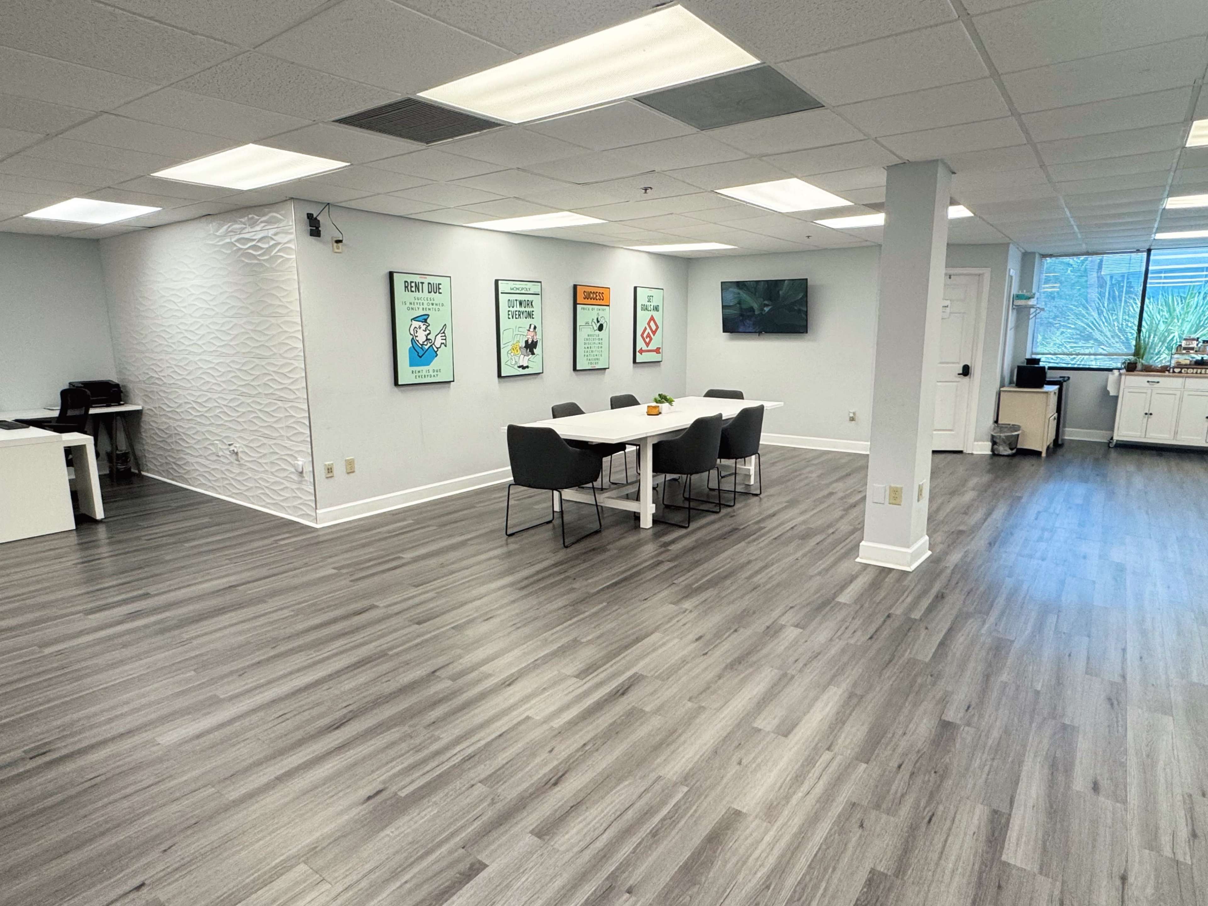 The image shows a well-lit office space with a central table surrounded by chairs, and walls adorned with framed posters.
