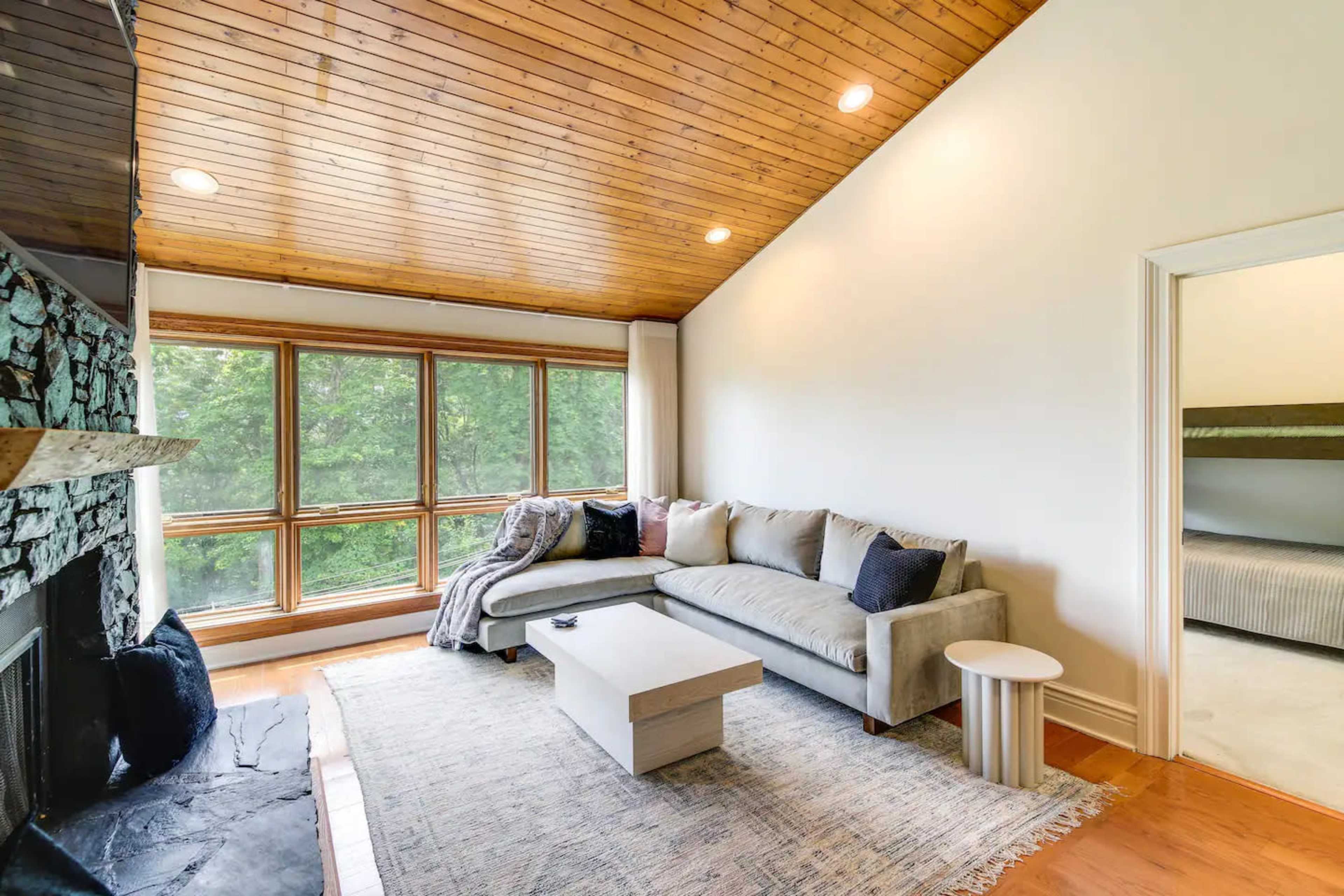 A modern living room features a sectional sofa, a small side table, a stone fireplace, and large windows that provide a view of the outdoors.