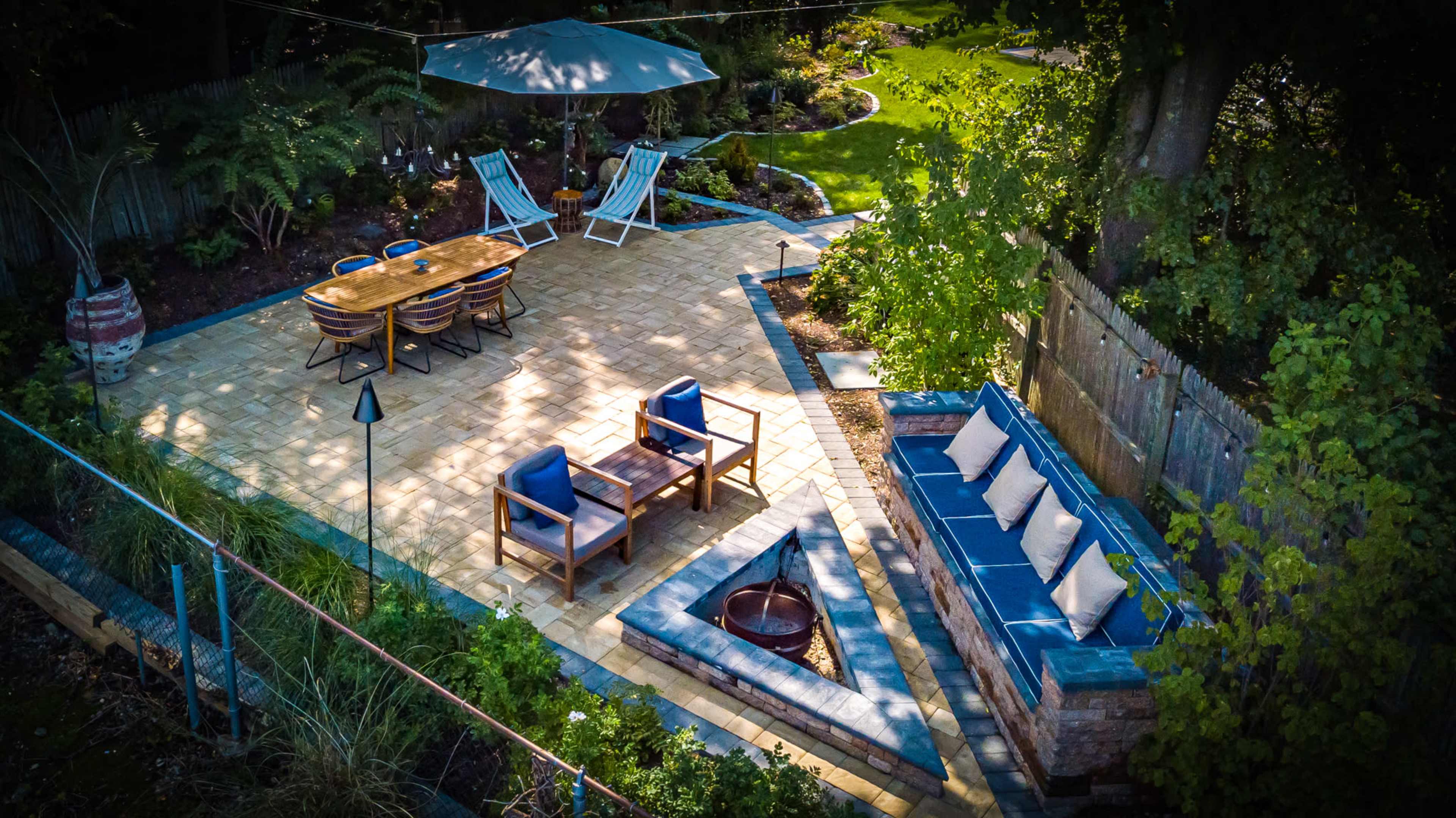 The image shows a landscaped patio area featuring outdoor seating, a dining table, and a fire pit surrounded by greenery.