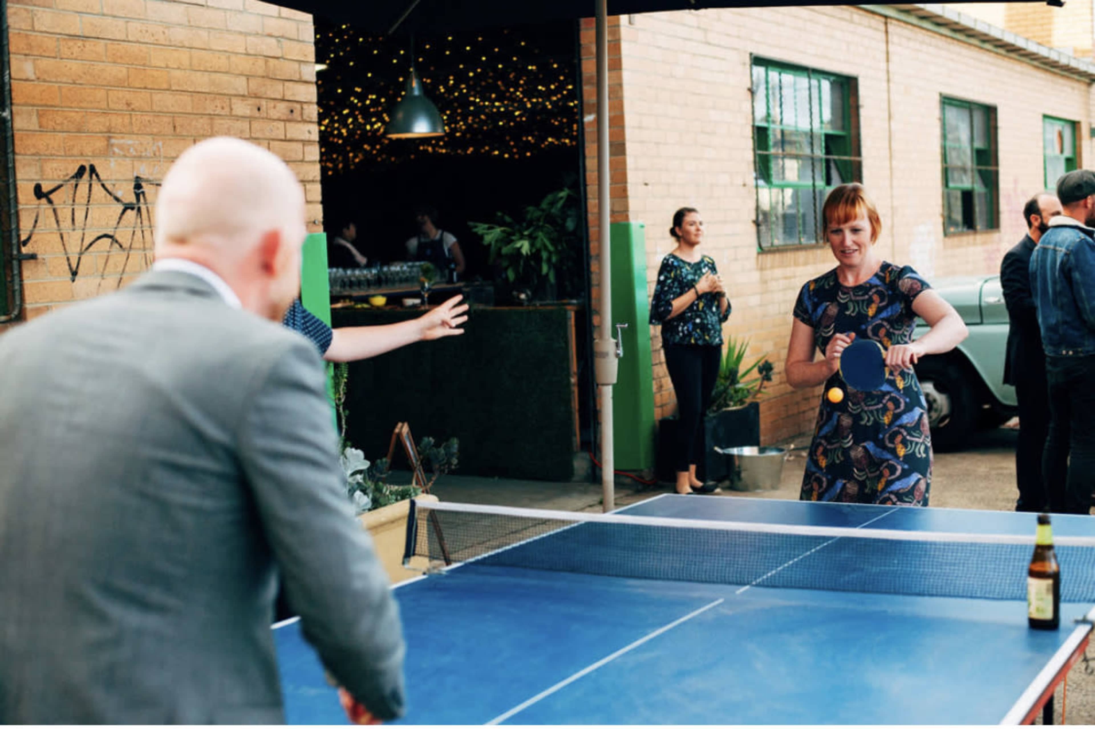 A woman in a patterned dress serves a ping pong ball across a blue table while a man in a suit prepares to return it in an outdoor setting.