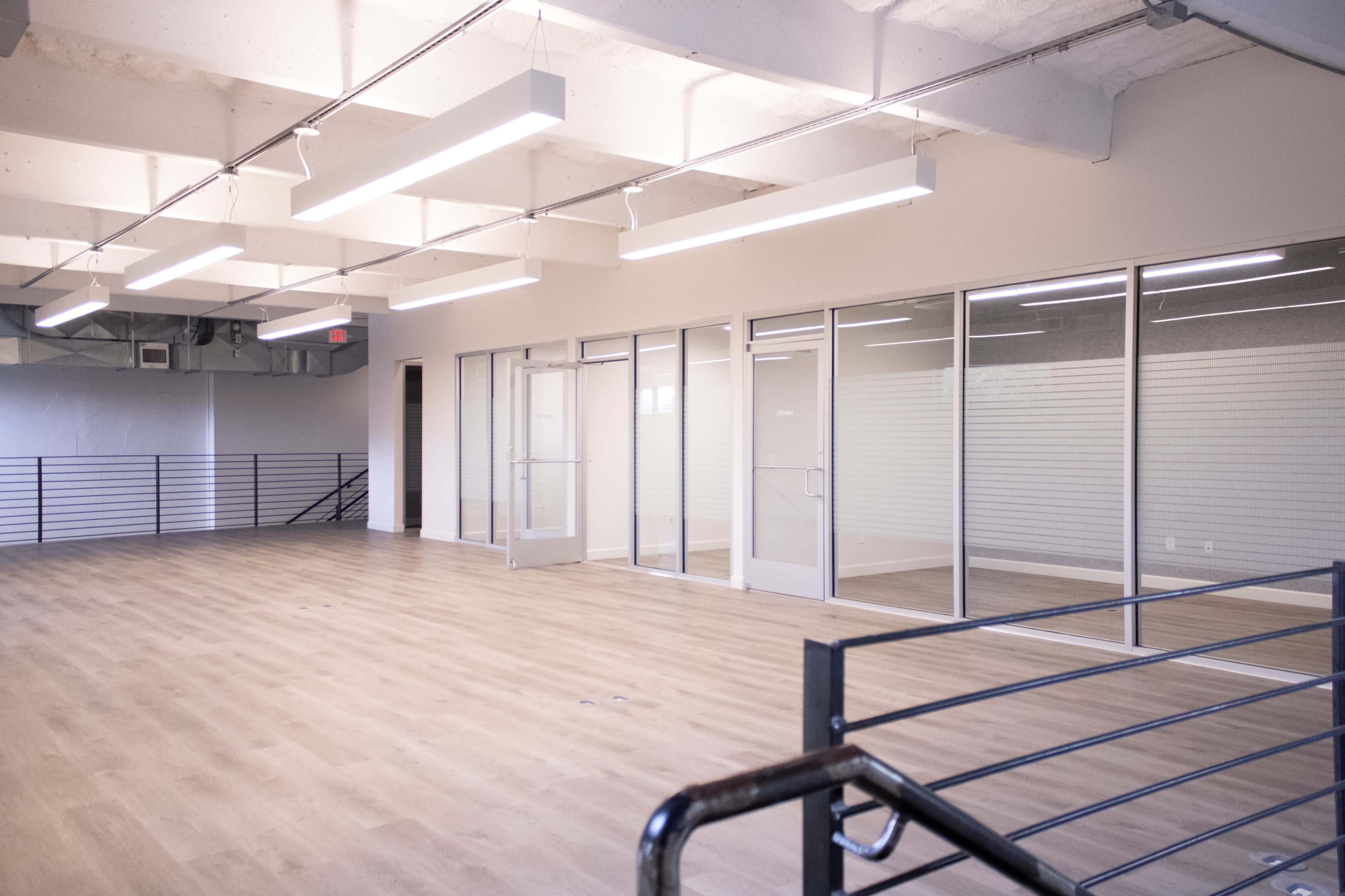The image shows a spacious, empty office area with large glass doors and windows, illuminated by long ceiling-mounted light fixtures.
