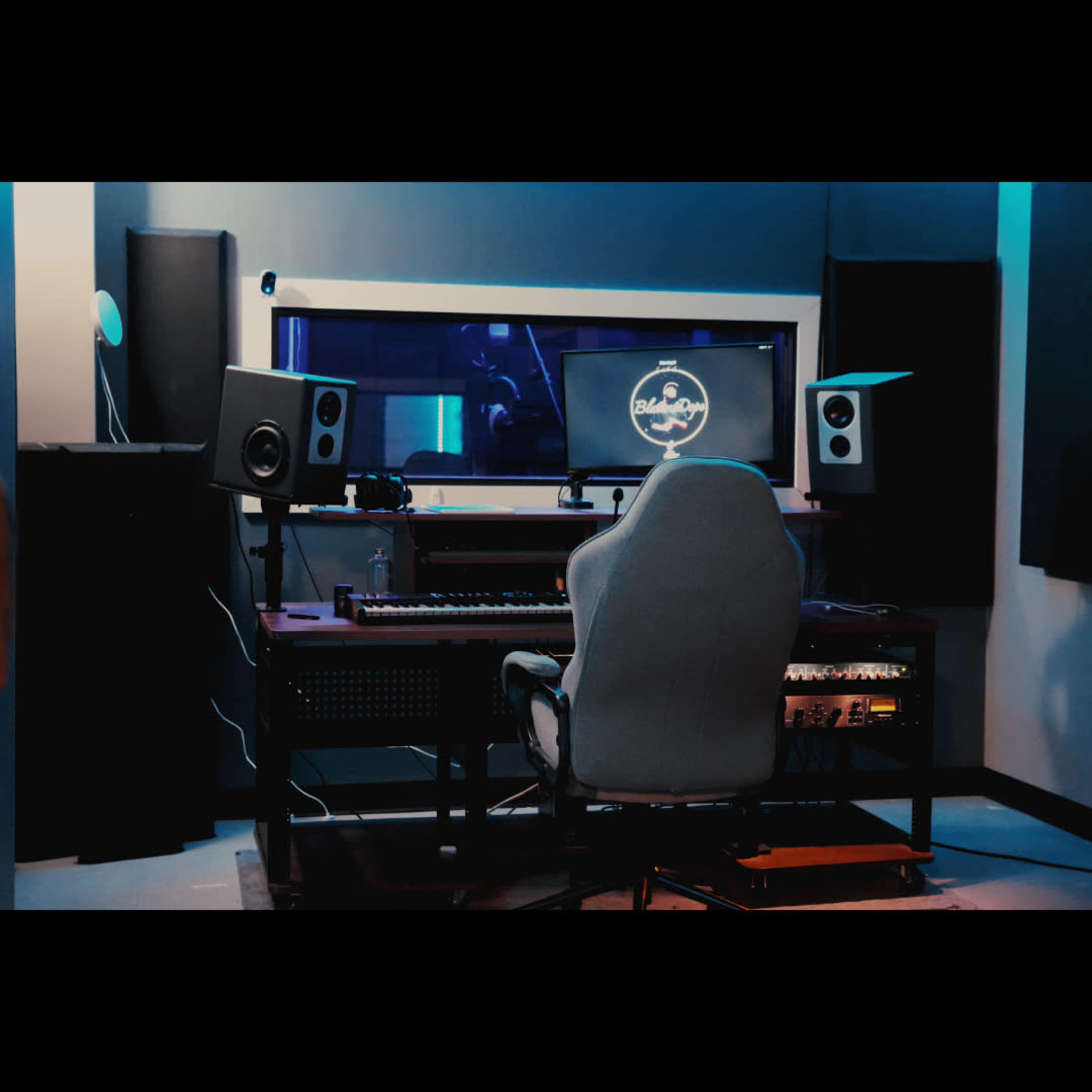 The image shows a music studio with a desk, a computer monitor, studio speakers, and an ergonomic chair set against a softly lit backdrop.