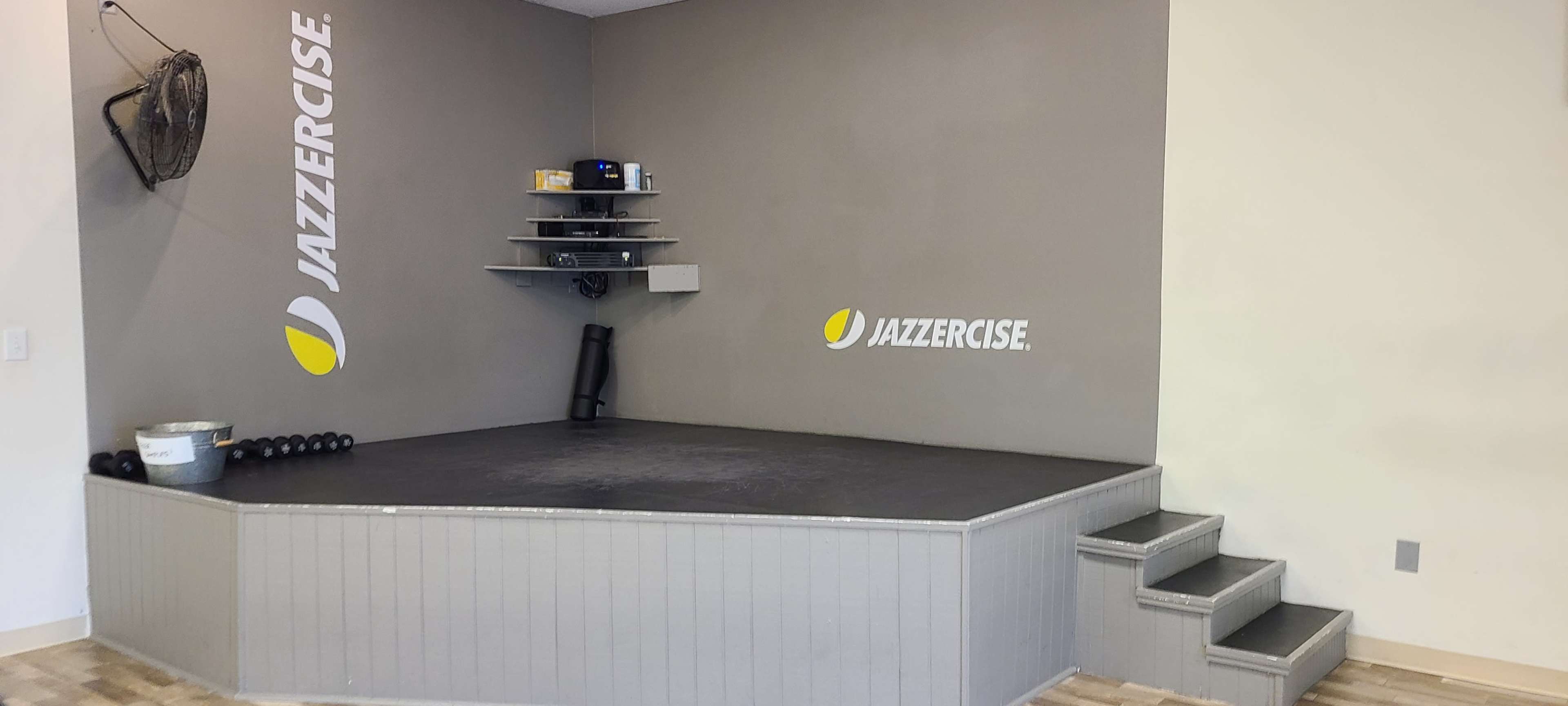 The image shows a raised, empty stage area with a gray wall featuring the Jazzercise logo and a set of shelves against it.