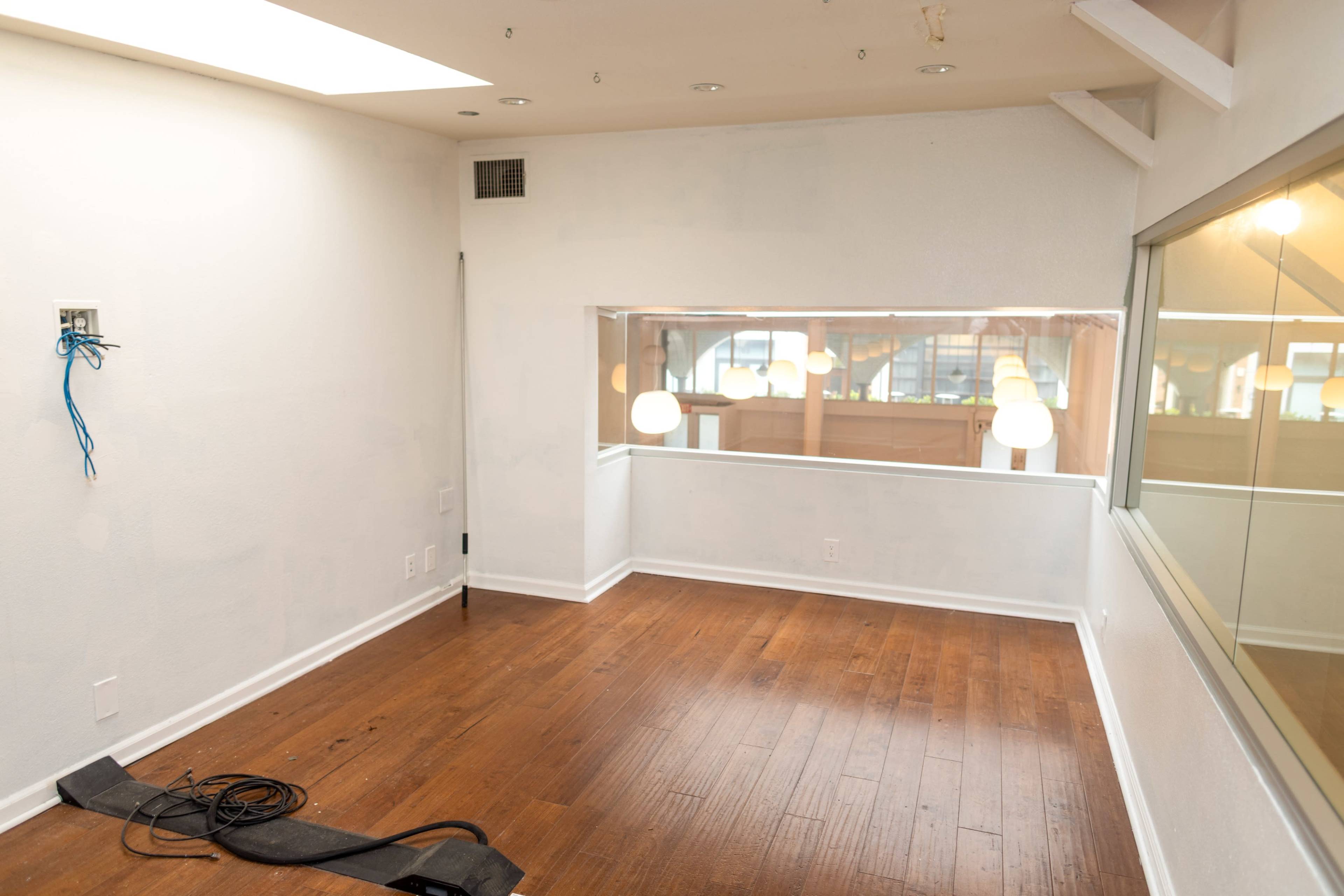 The image shows a partially empty room with hardwood flooring, a large window, and bare walls in a well-lit space.