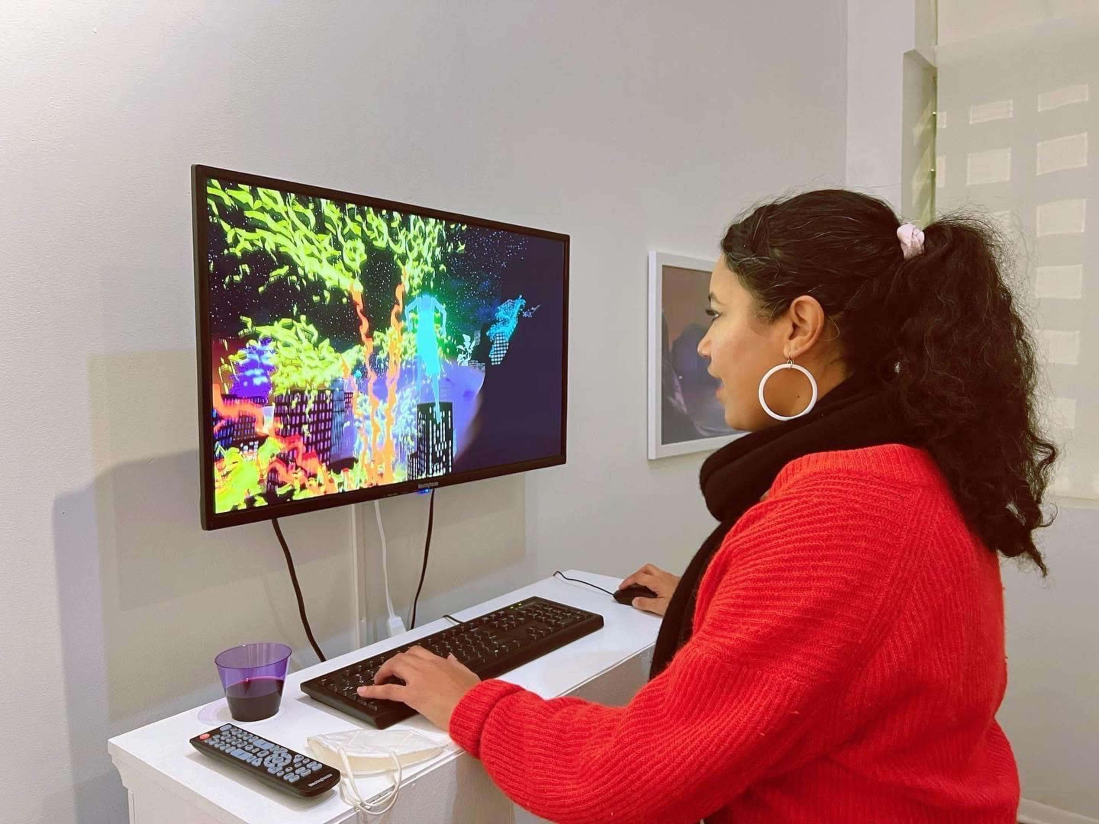 A woman in a red sweater is seated at a computer, interacting with a colorful digital display in an art gallery.
