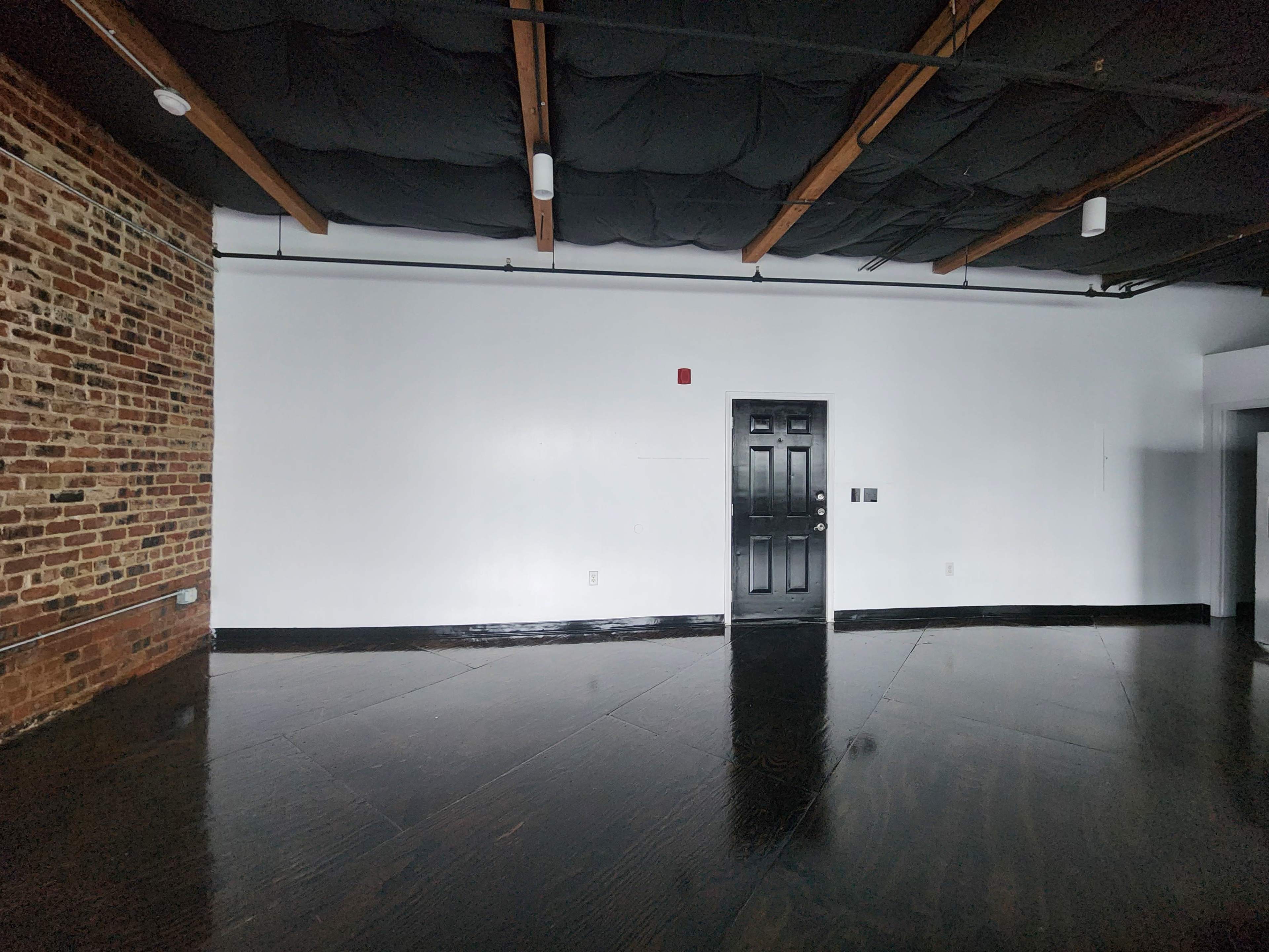 The image shows a spacious room with a black door, exposed brick wall, and dark wooden flooring.