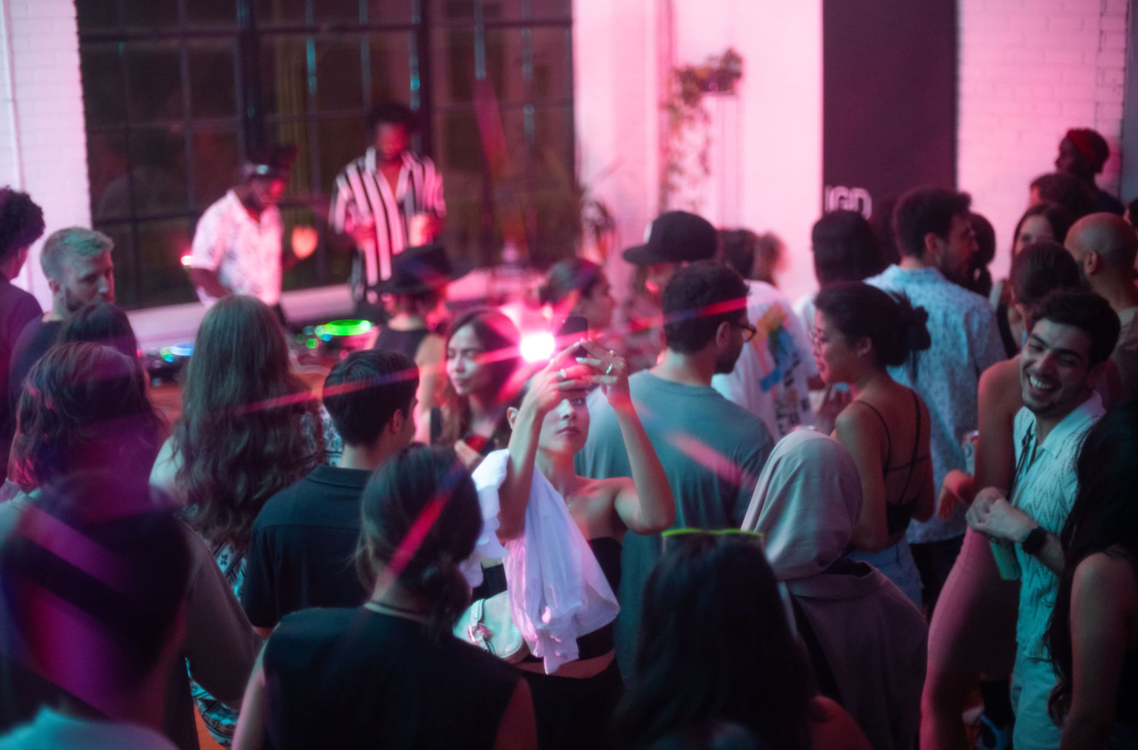 A crowded indoor party scene features people socializing and dancing under vibrant pink and purple lights, with a DJ setup in the background.