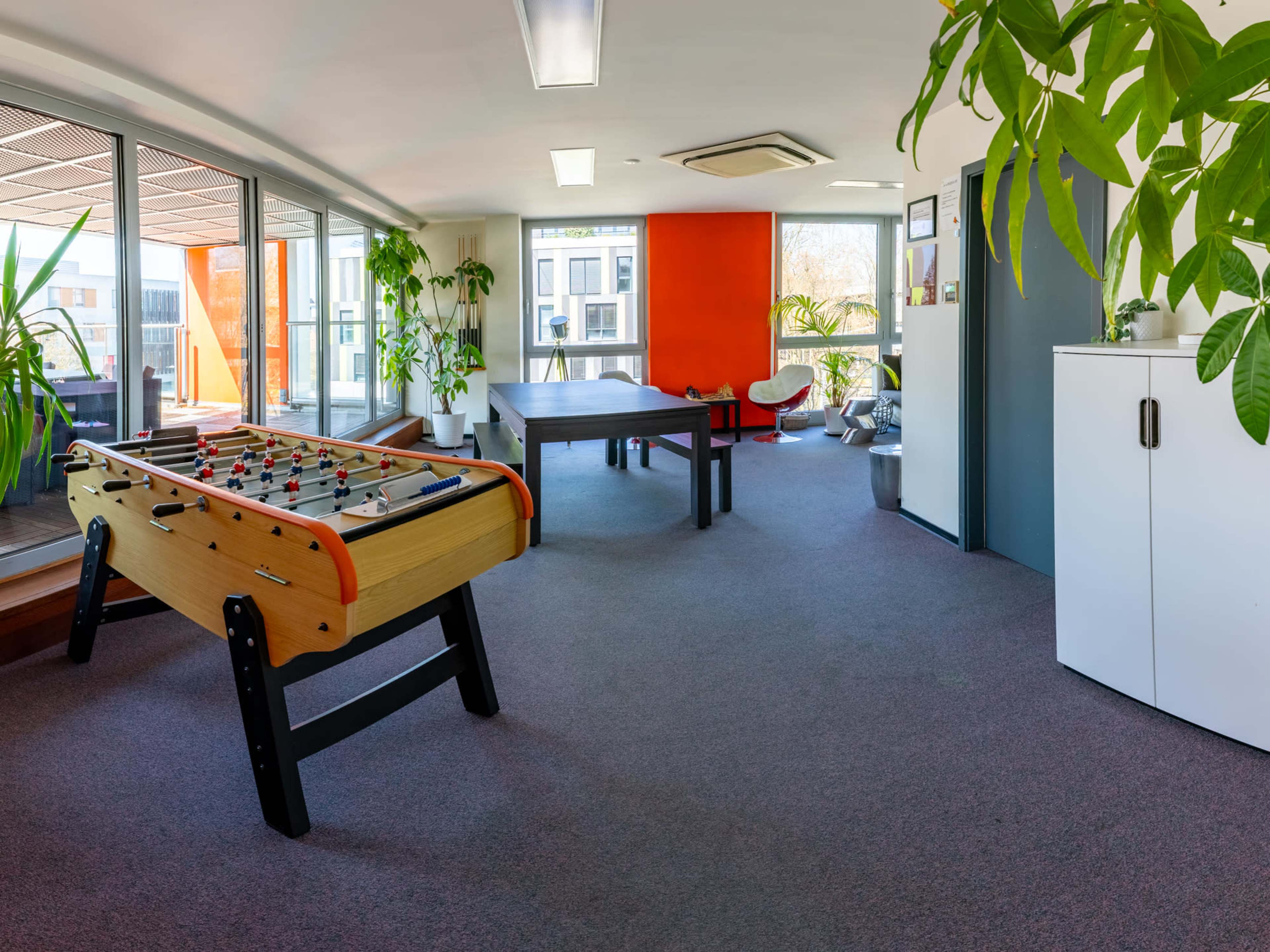 The image shows a modern indoor recreational area featuring a foosball table, a ping pong table, and several potted plants.
