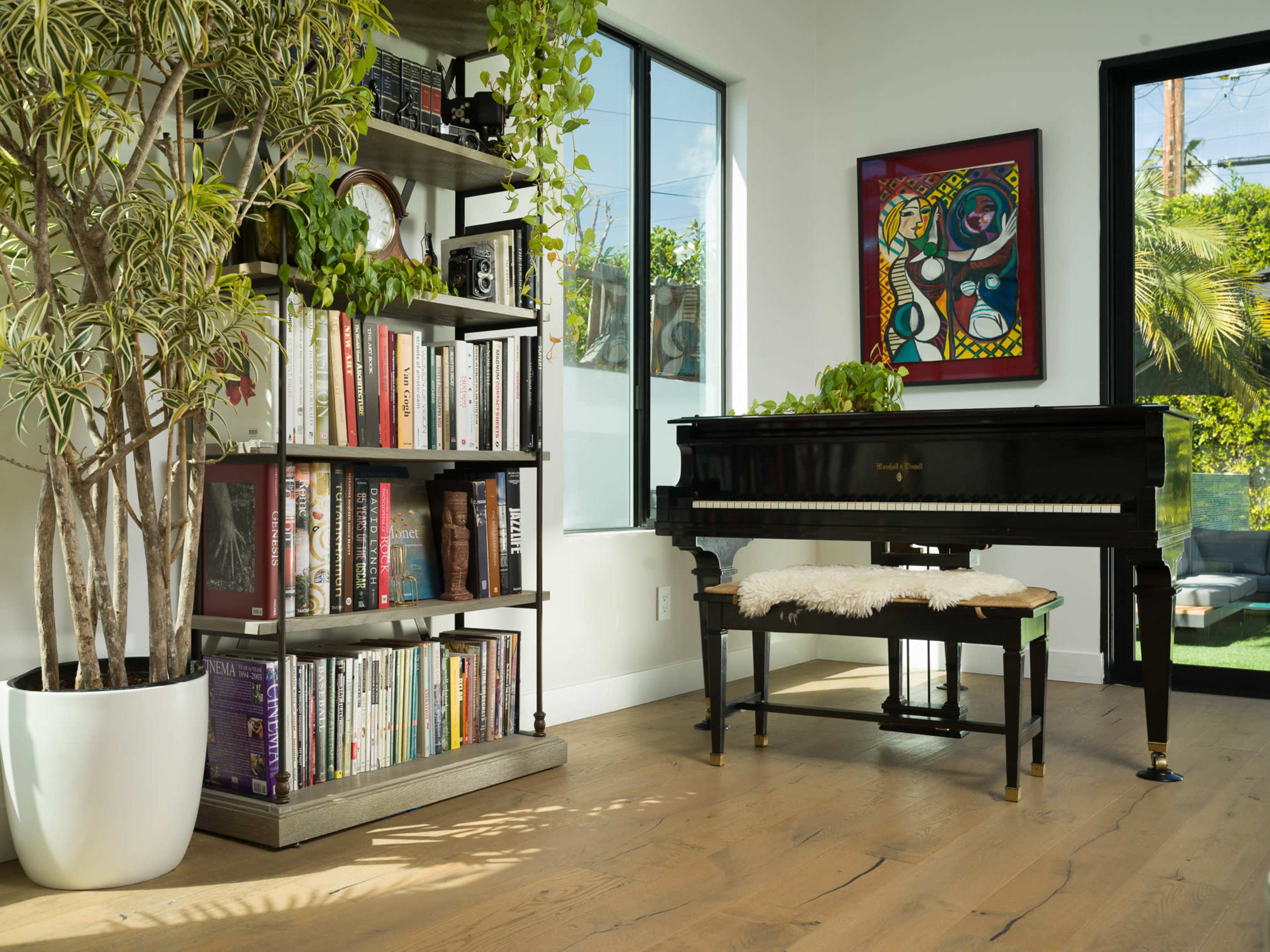 A black grand piano is positioned beside a bookshelf filled with books and surrounded by plants in a well-lit room with large windows.