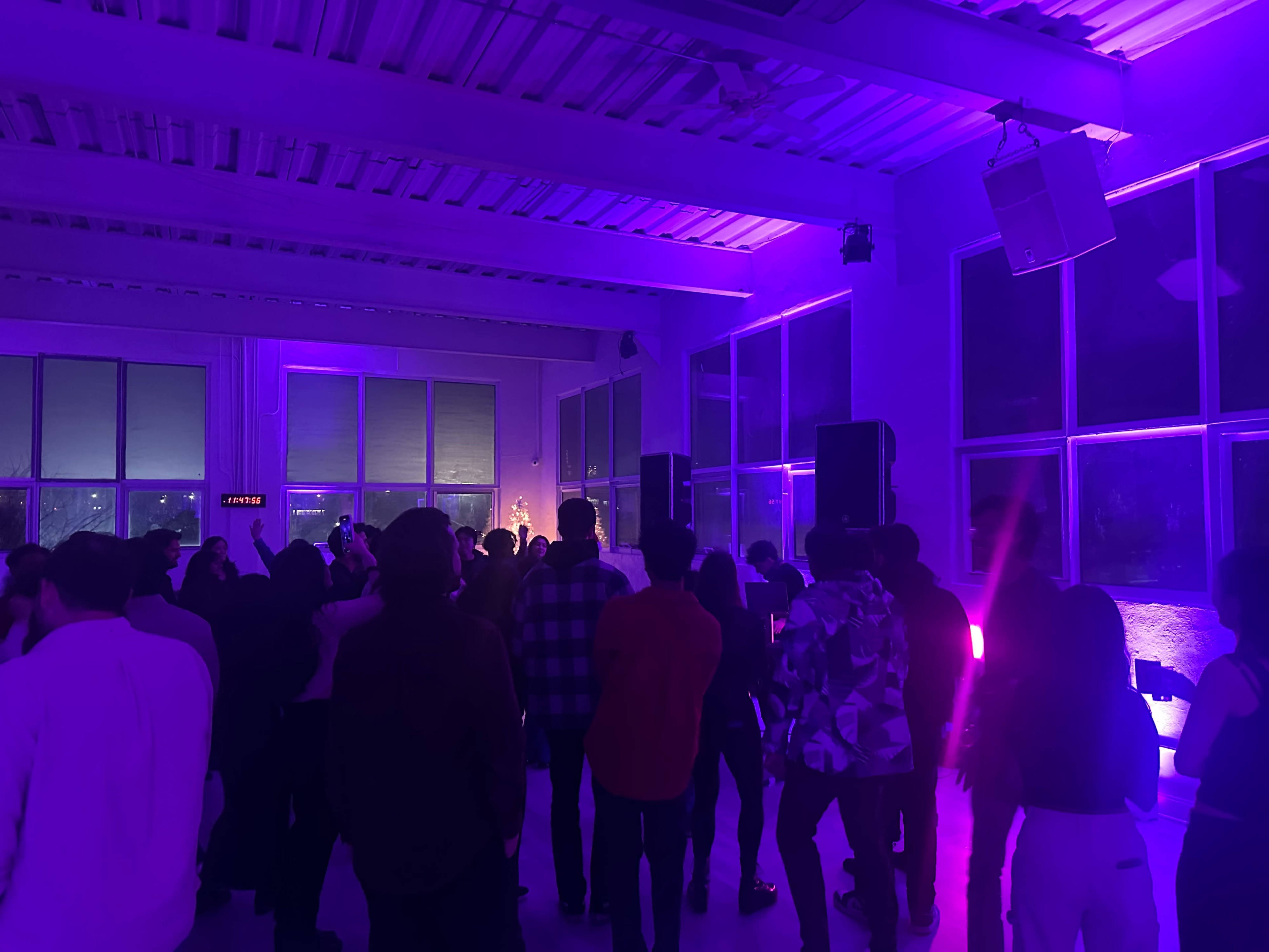 A crowd of people dances in a brightly lit room adorned with purple lighting and large windows.