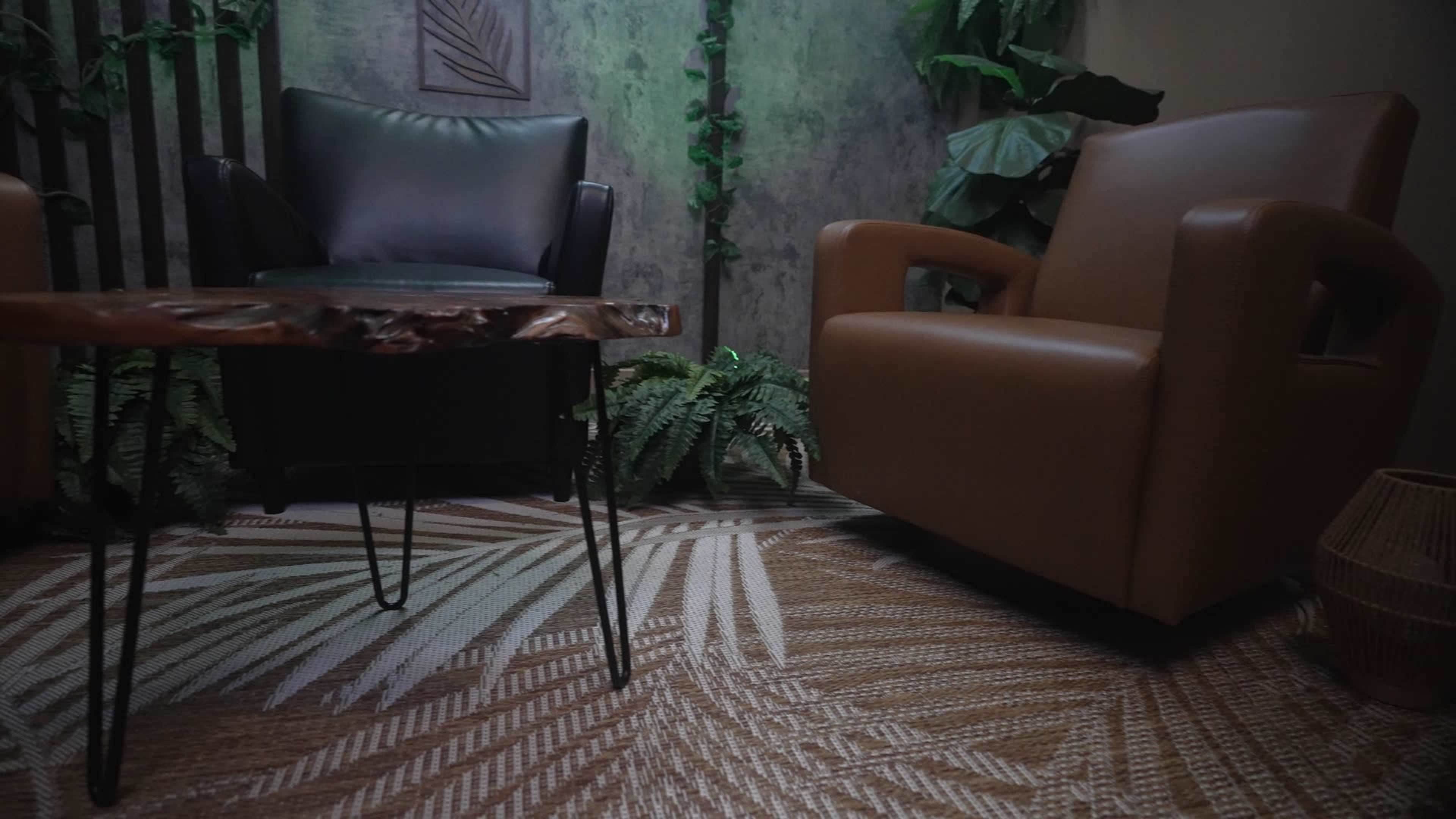 The image shows two armchairs, one black and one brown, facing each other across a wooden table with slender legs, surrounded by greenery and textured walls.
