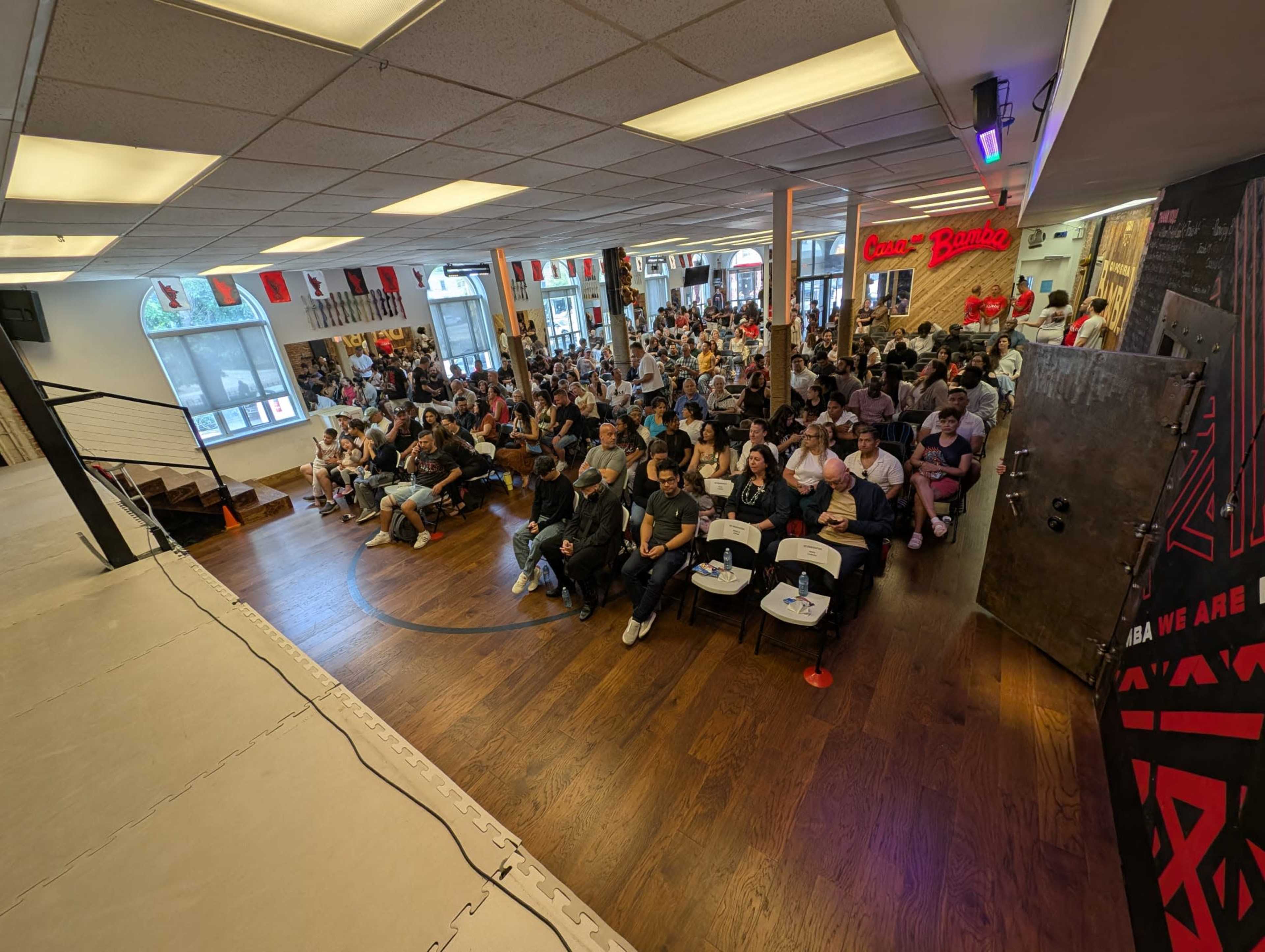 A large crowd of people sits in rows of chairs facing a stage in a brightly lit indoor venue decorated with flags and banners.